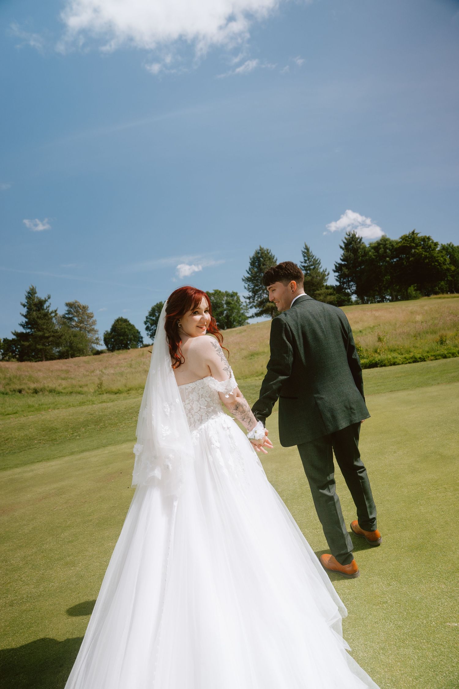 Bride in white gown and veil holds groom's hand walking on green lawn under blue sky.