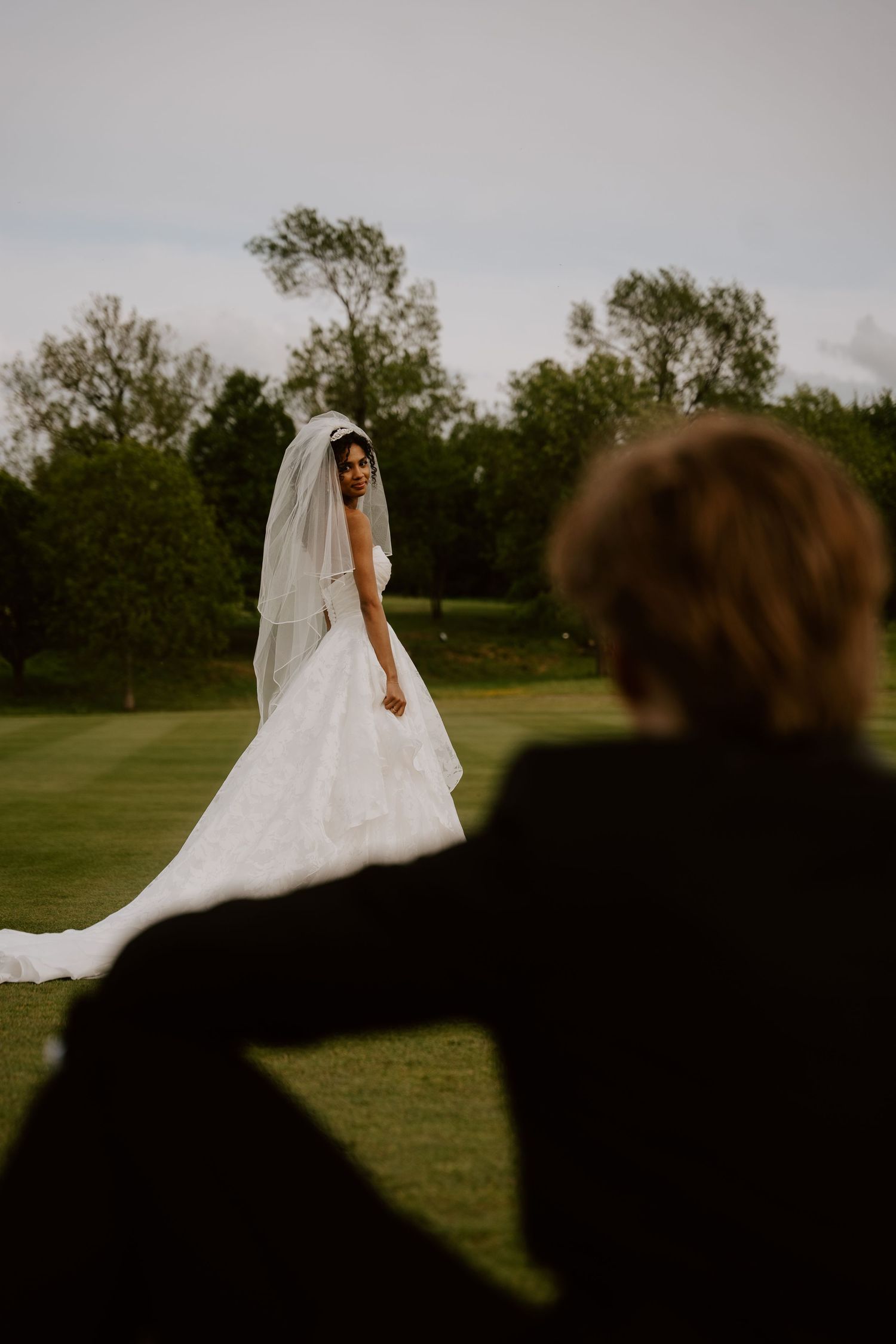 Bride in white gown and veil stands on green lawn as groom watches from foreground at outdoor wedding.