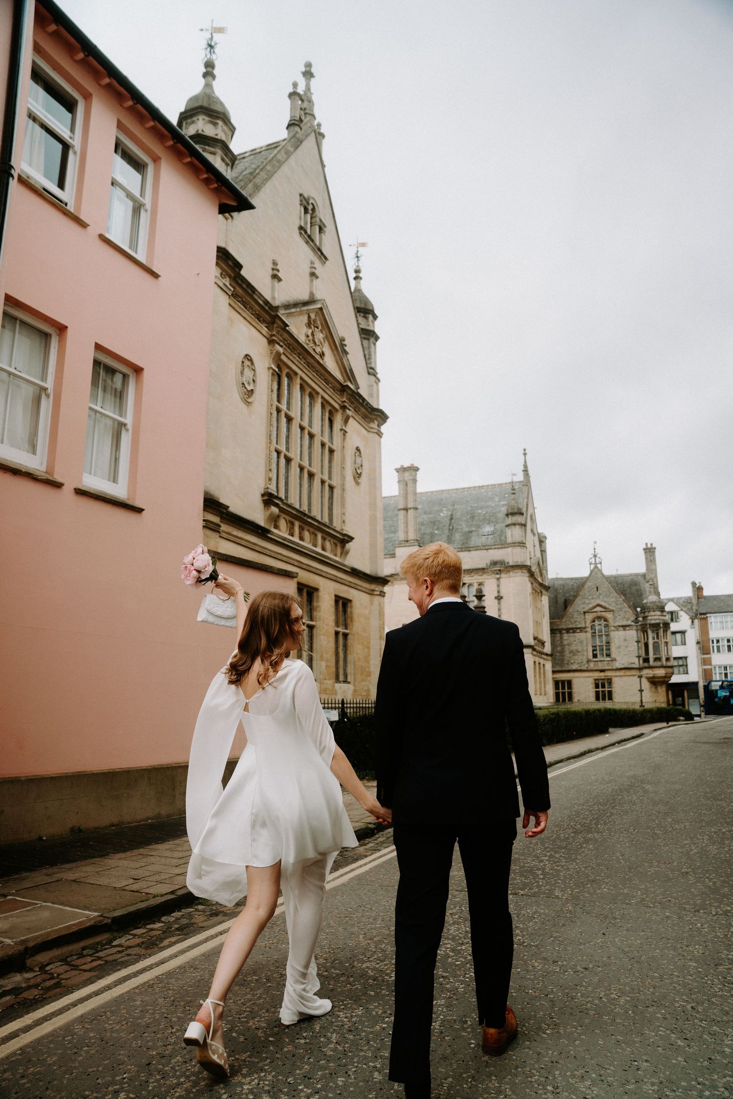 Bride and groom walking hand in hand down a historic street, bride in short white dress holding pink bouquet.
