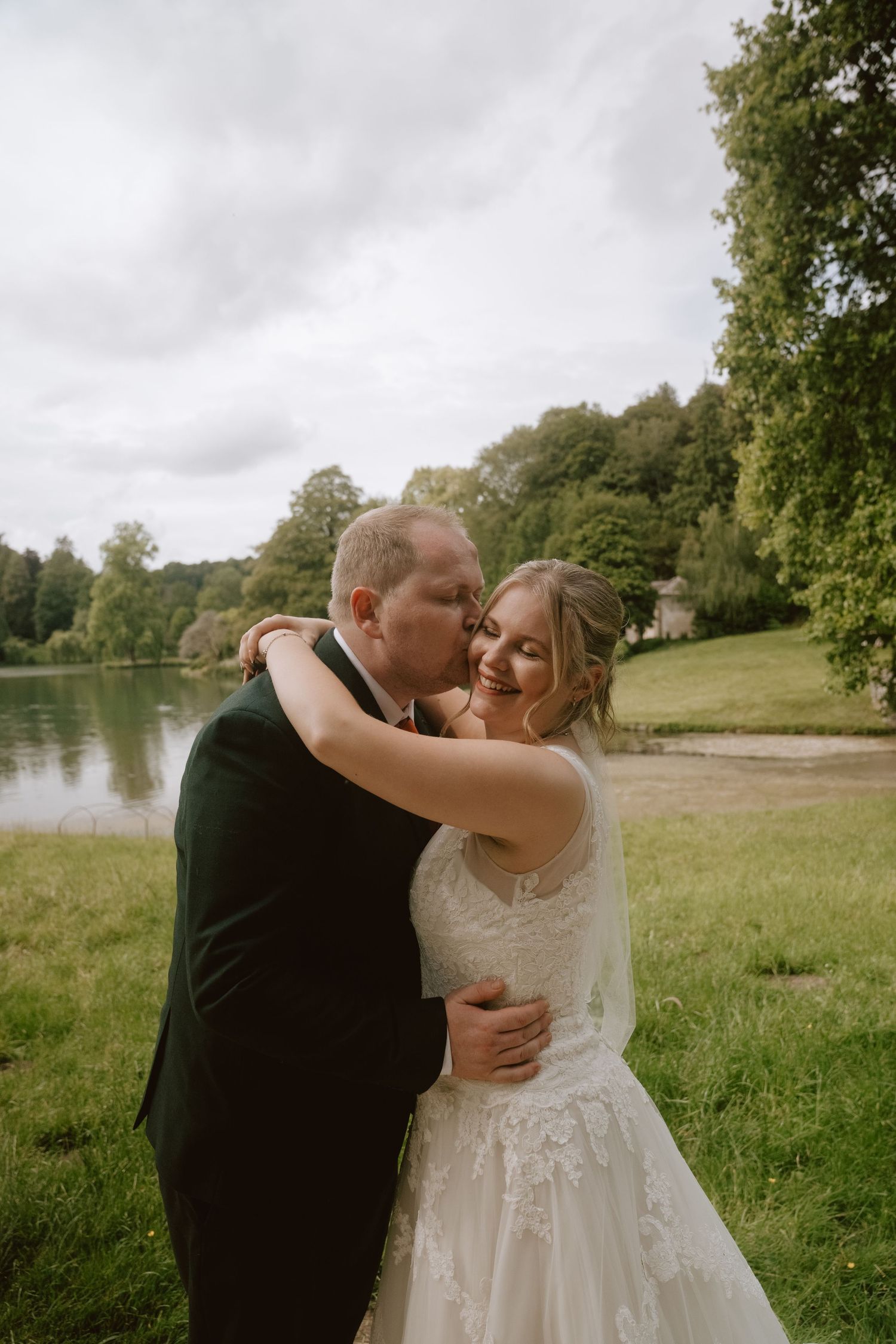 A groom kisses his bride on the cheek as they embrace outdoors near a lake surrounded by lush greenery on their wedding day.