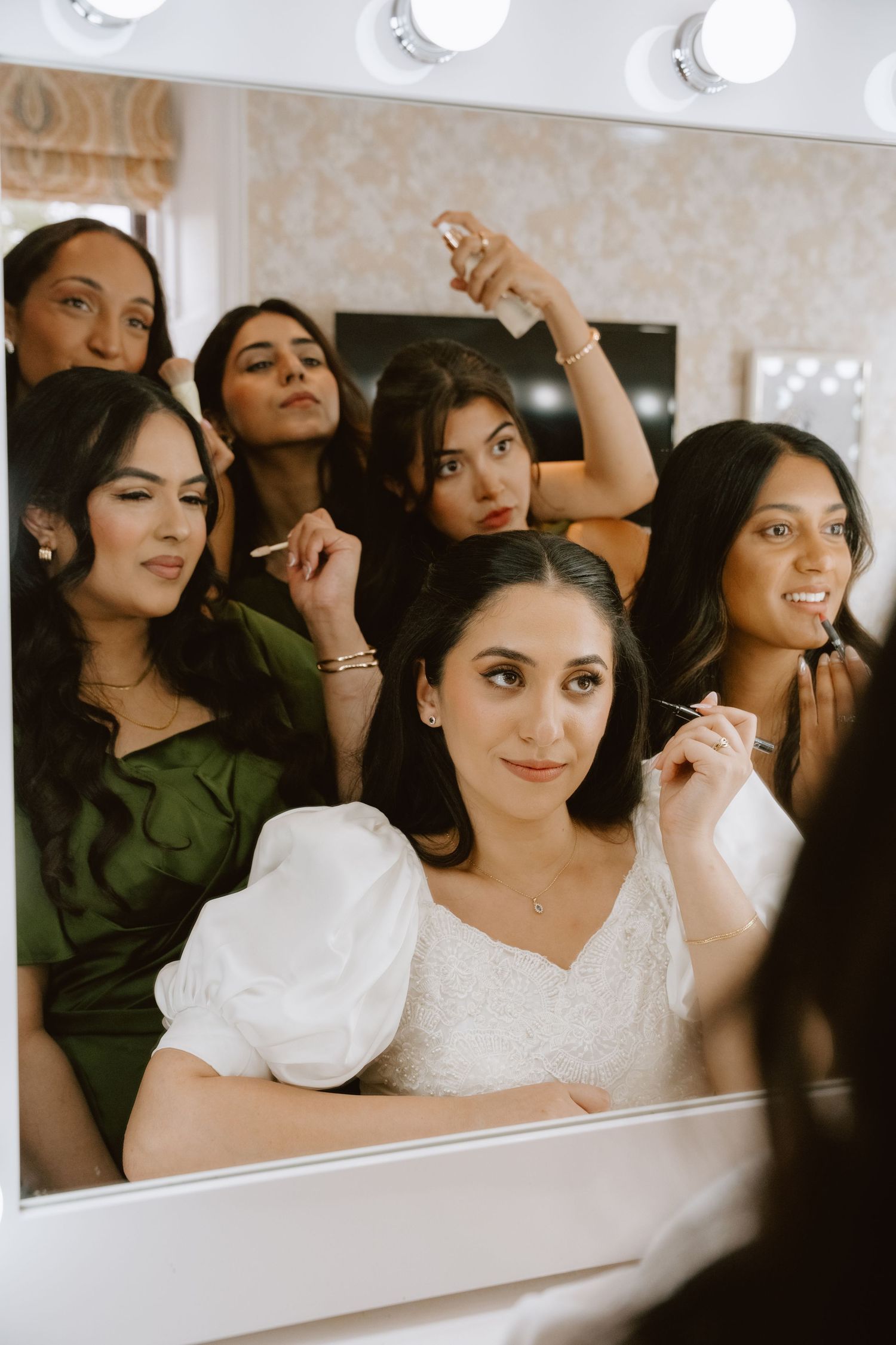 Bride in white dress with bridesmaids in green reflected in a Hollywood mirror with lights during wedding preparations.