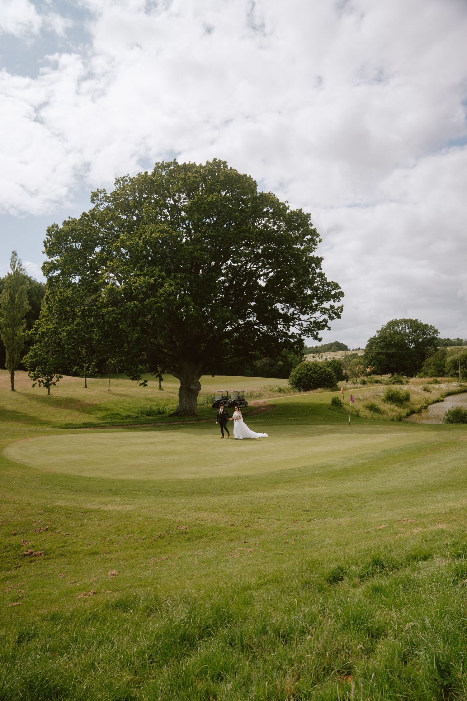 A bride and groom stand beneath a large oak tree on a lush green golf course under a partly cloudy sky.