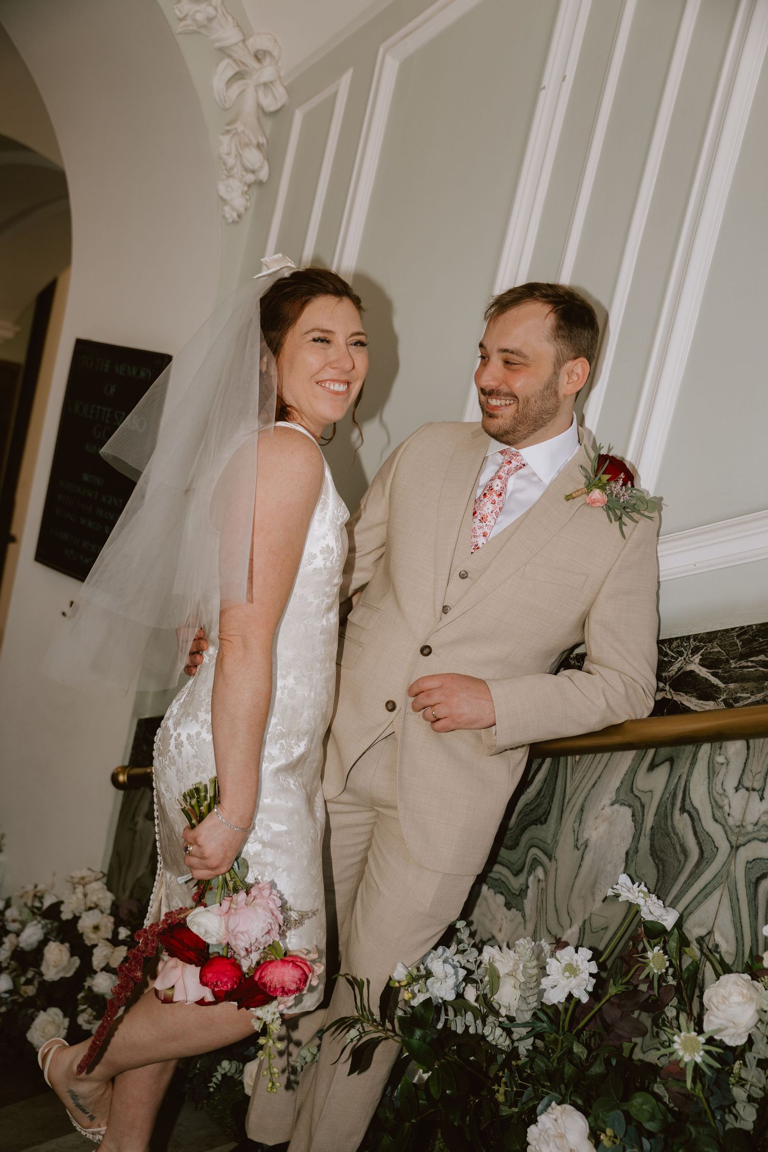 Bride in white mini dress and groom in beige suit smiling on decorated staircase with floral arrangements.