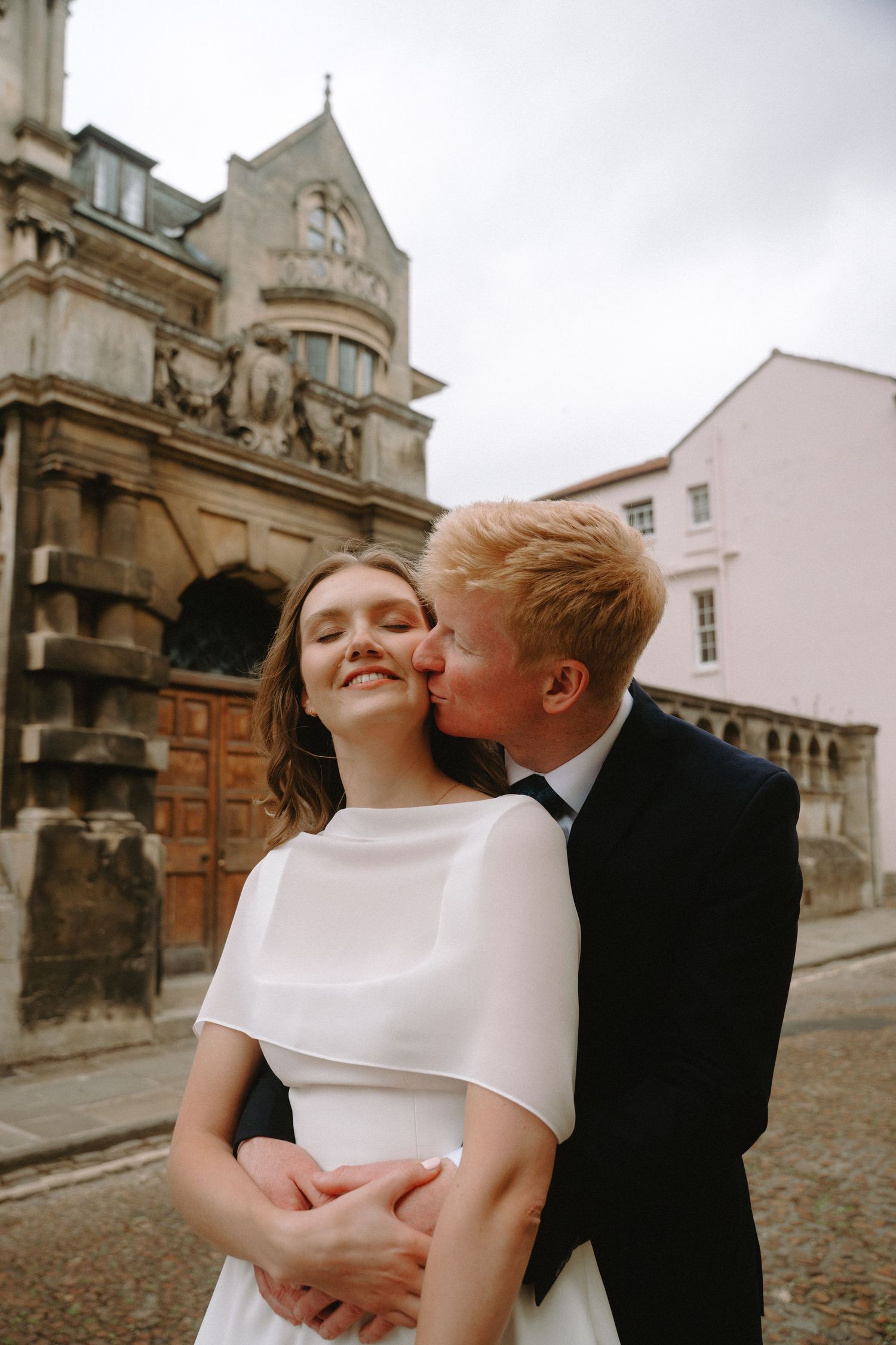 A groom with red hair kisses his smiling bride in a white dress on a cobblestone street with historic buildings.