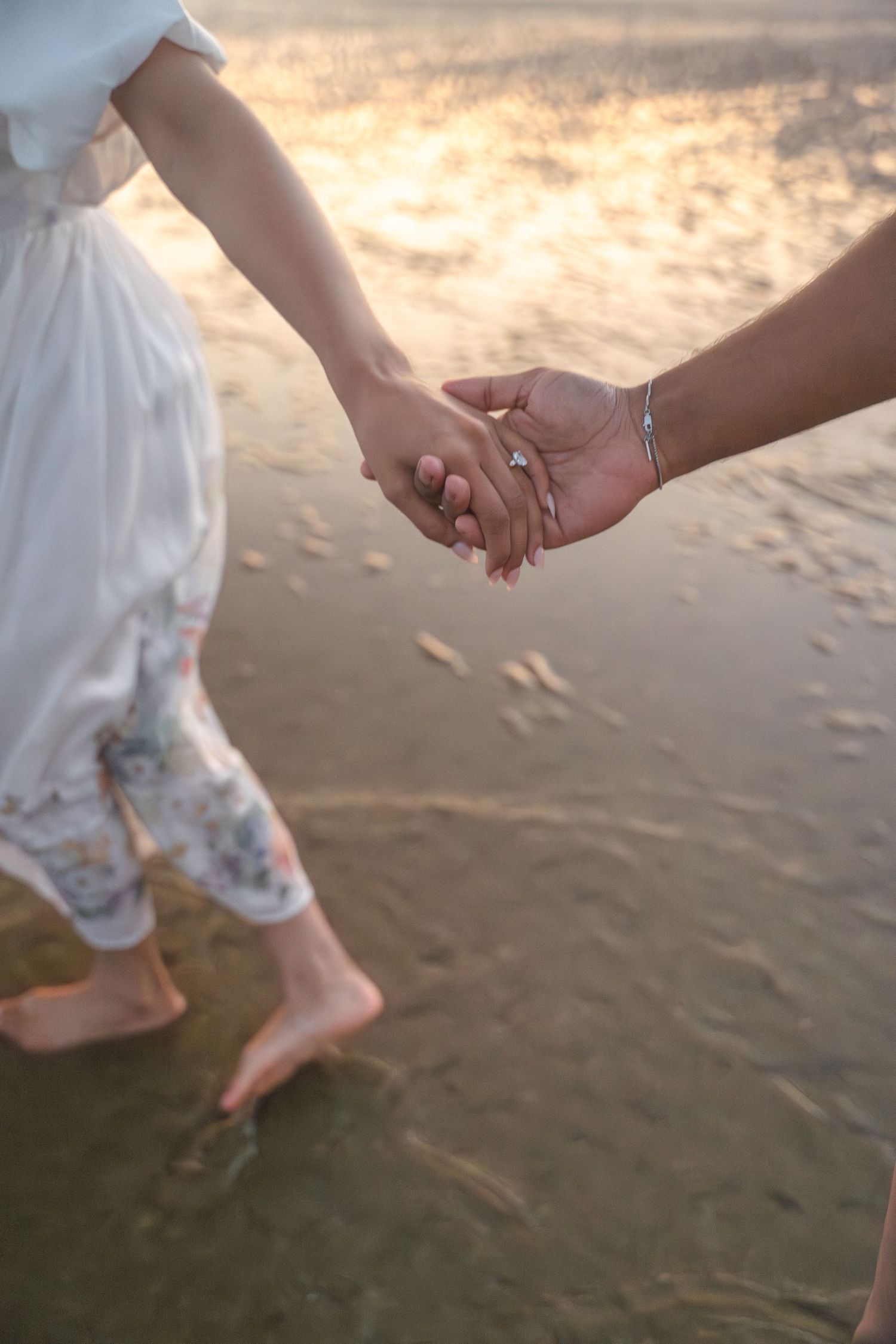 Couple holding hands while walking barefoot on wet beach sand at sunset.