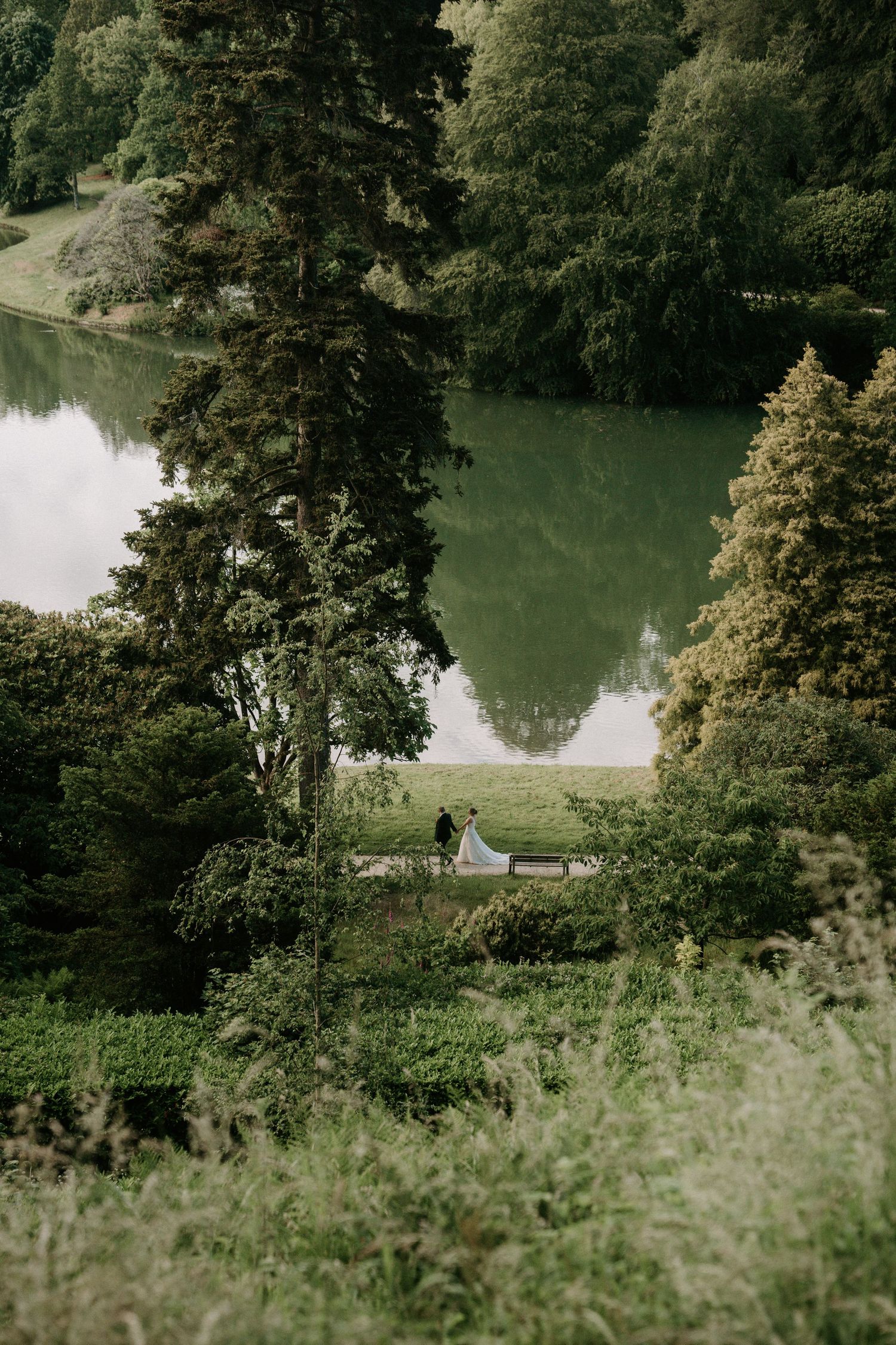 Aerial view of a couple near a serene green lake surrounded by lush trees in a scenic garden landscape.