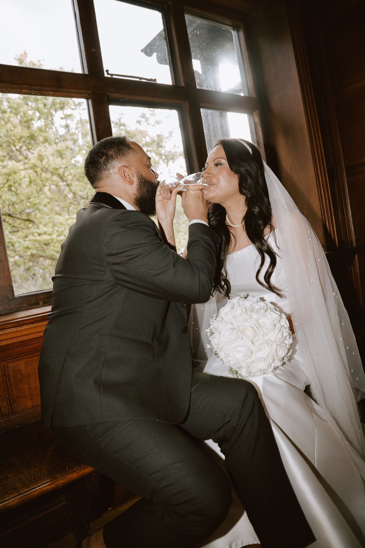 Bride and groom sharing a champagne toast by a sunlit window, bride holding white rose bouquet in wedding attire.