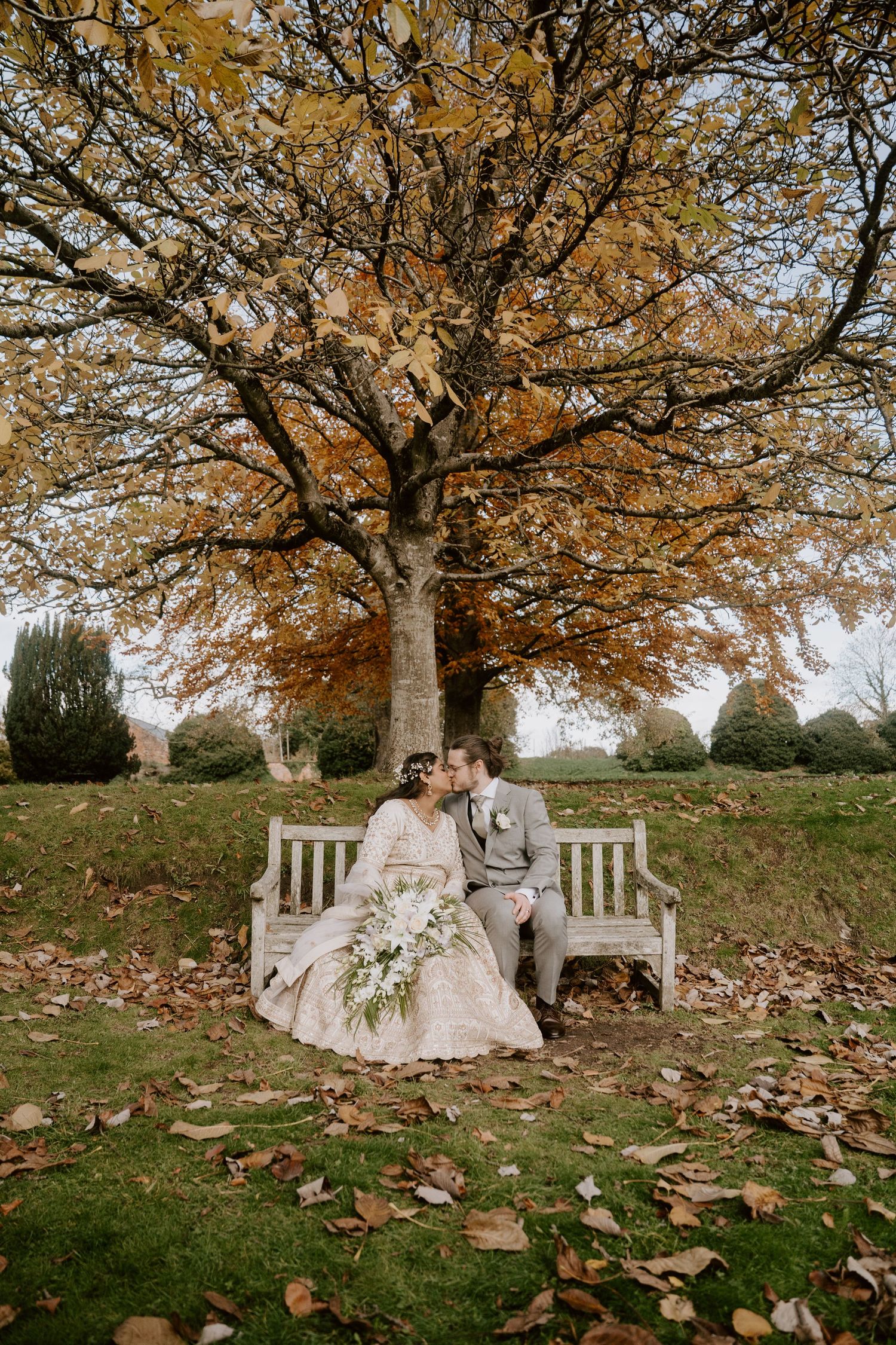 Romantic autumn wedding scene with couple sitting on white bench under golden tree with fallen leaves.