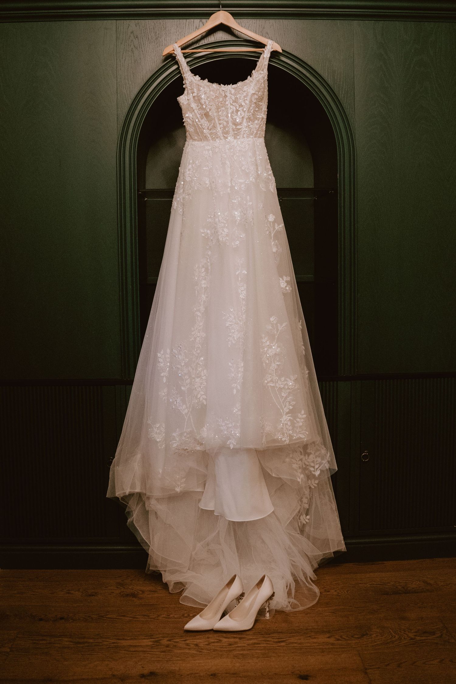 A white lace wedding dress hangs elegantly against a dark wall with hardwood flooring visible below.
