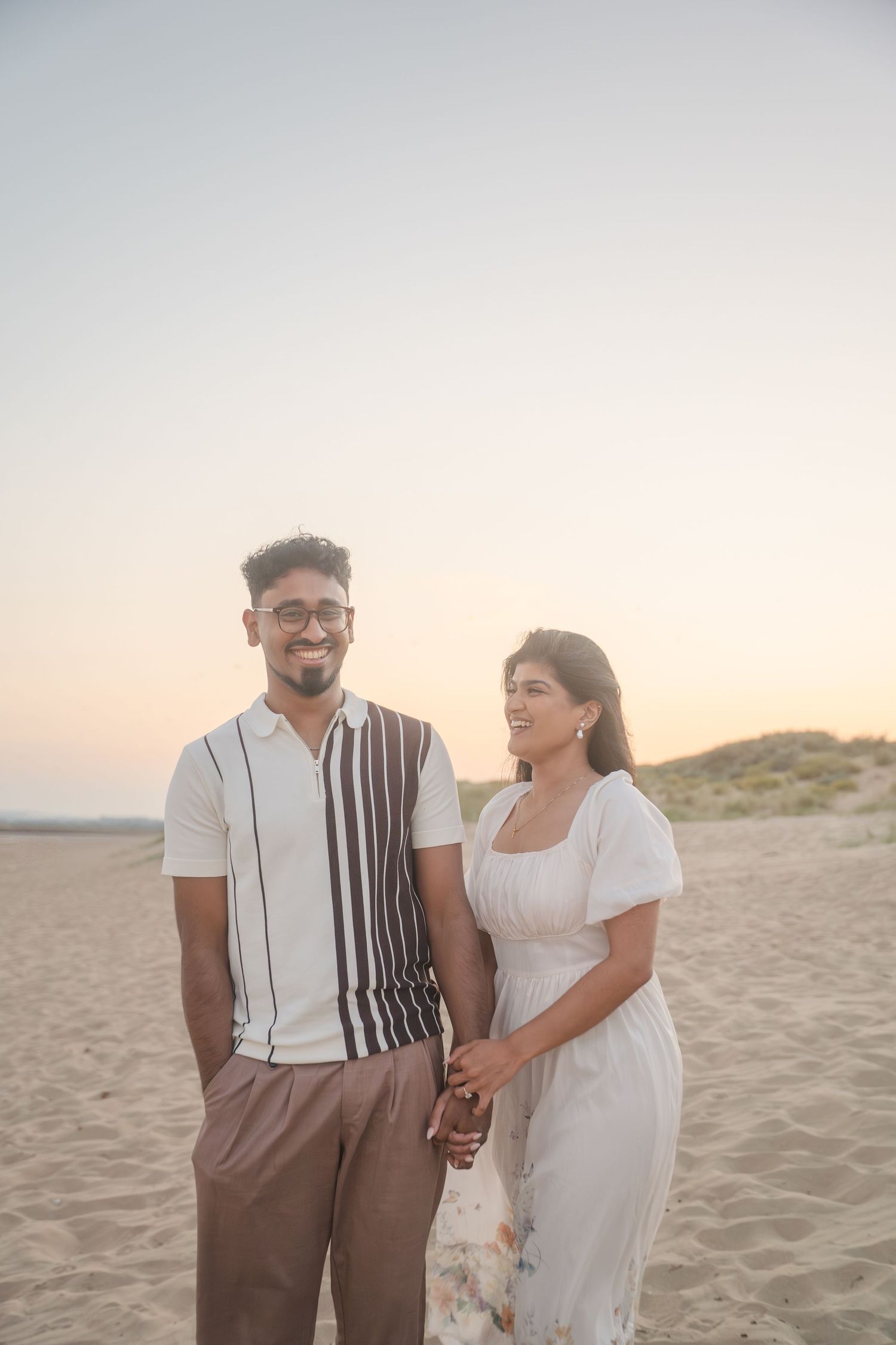 A couple in coordinated white outfits holds hands while standing on a sandy beach at sunset.