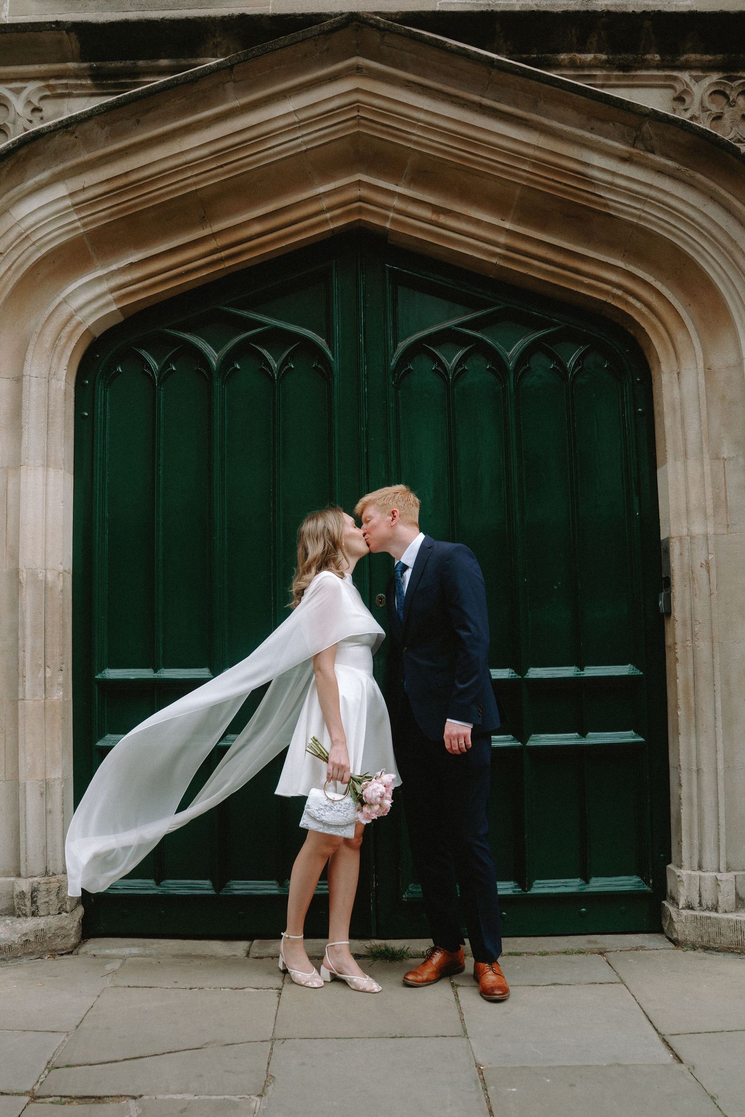 Bride and groom share a kiss in front of a grand green Gothic door, bride wearing a short white dress with flowing cape.