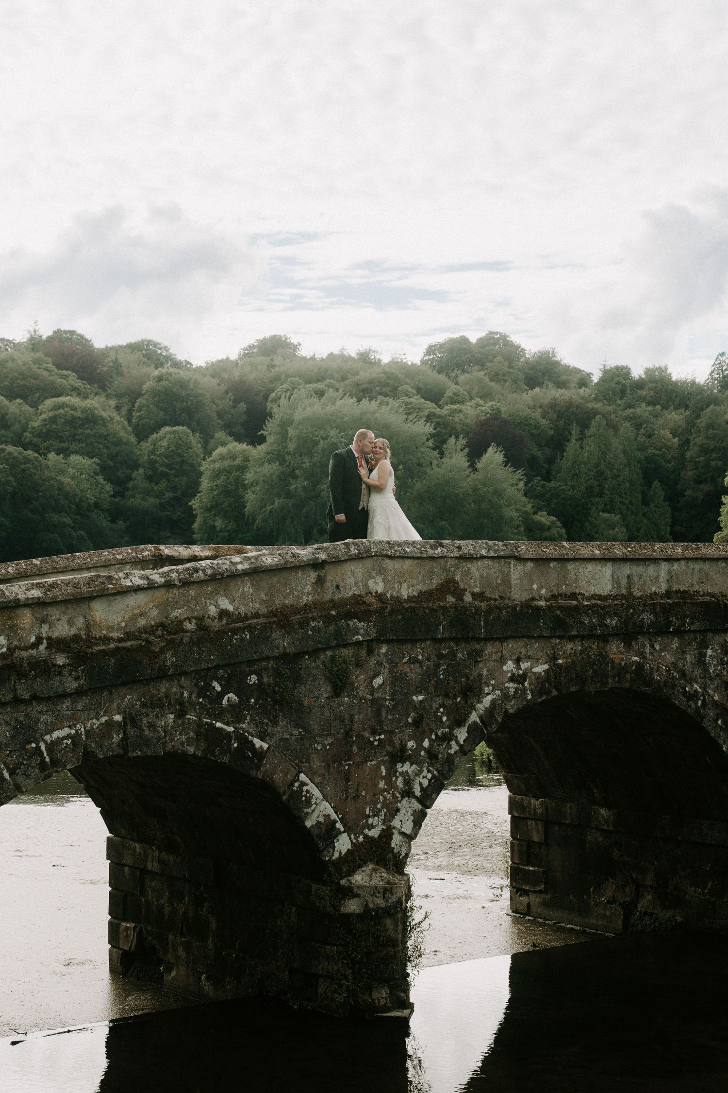 Bride and groom embracing on a stone arch bridge surrounded by lush green trees on their wedding day.