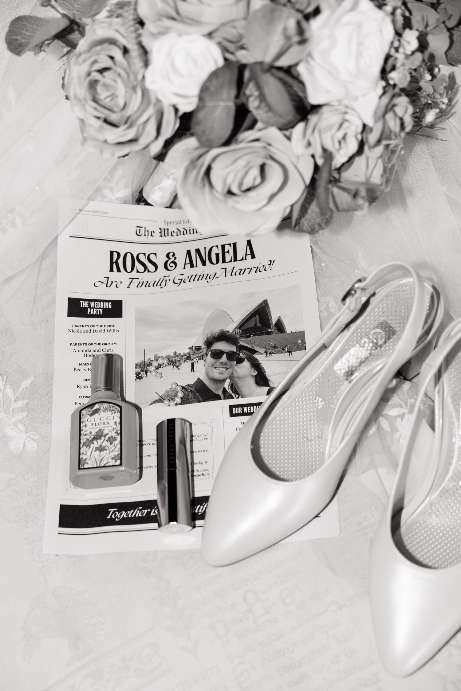 Black and white wedding flat lay featuring Ross & Angela newspaper, bridal heels, perfume bottles, and a rose bouquet.