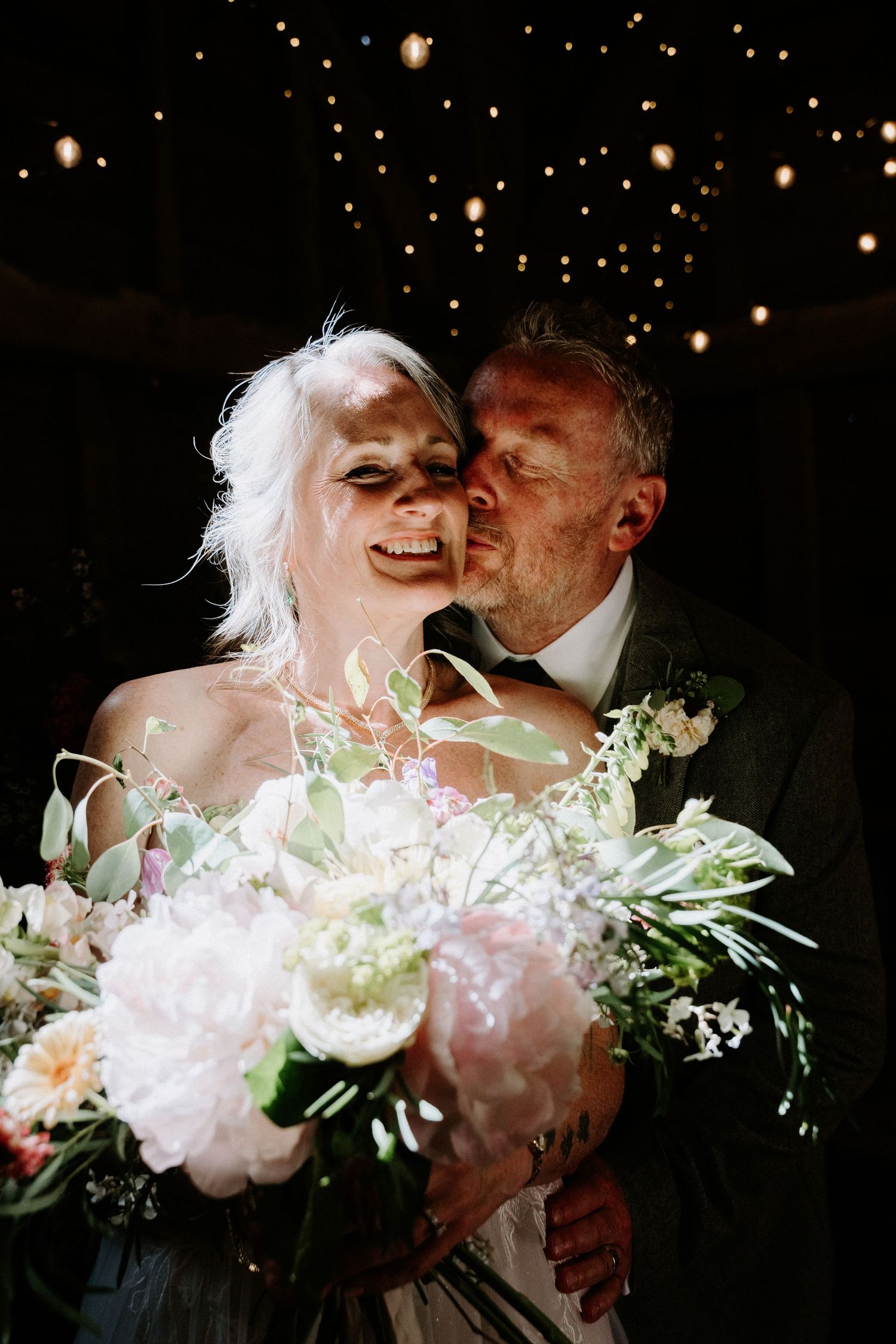 A joyful bride holding a lush floral bouquet receives a kiss from the groom under twinkling fairy lights.