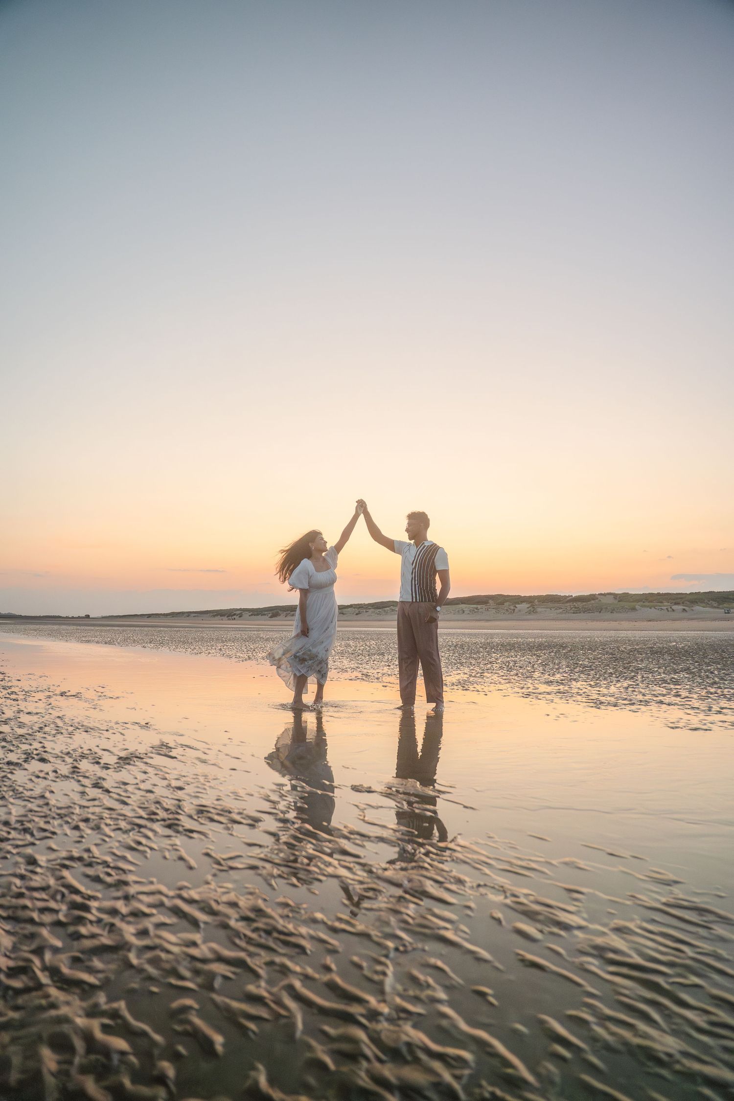 Two people hold hands and dance in the shallow water at sunset, creating reflections on the wet sand.