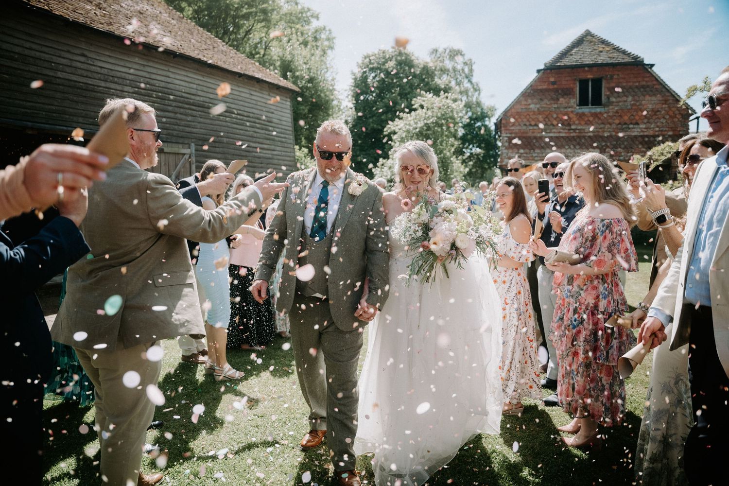 Bride and groom walk through confetti shower surrounded by cheering wedding guests outside a rustic barn venue.