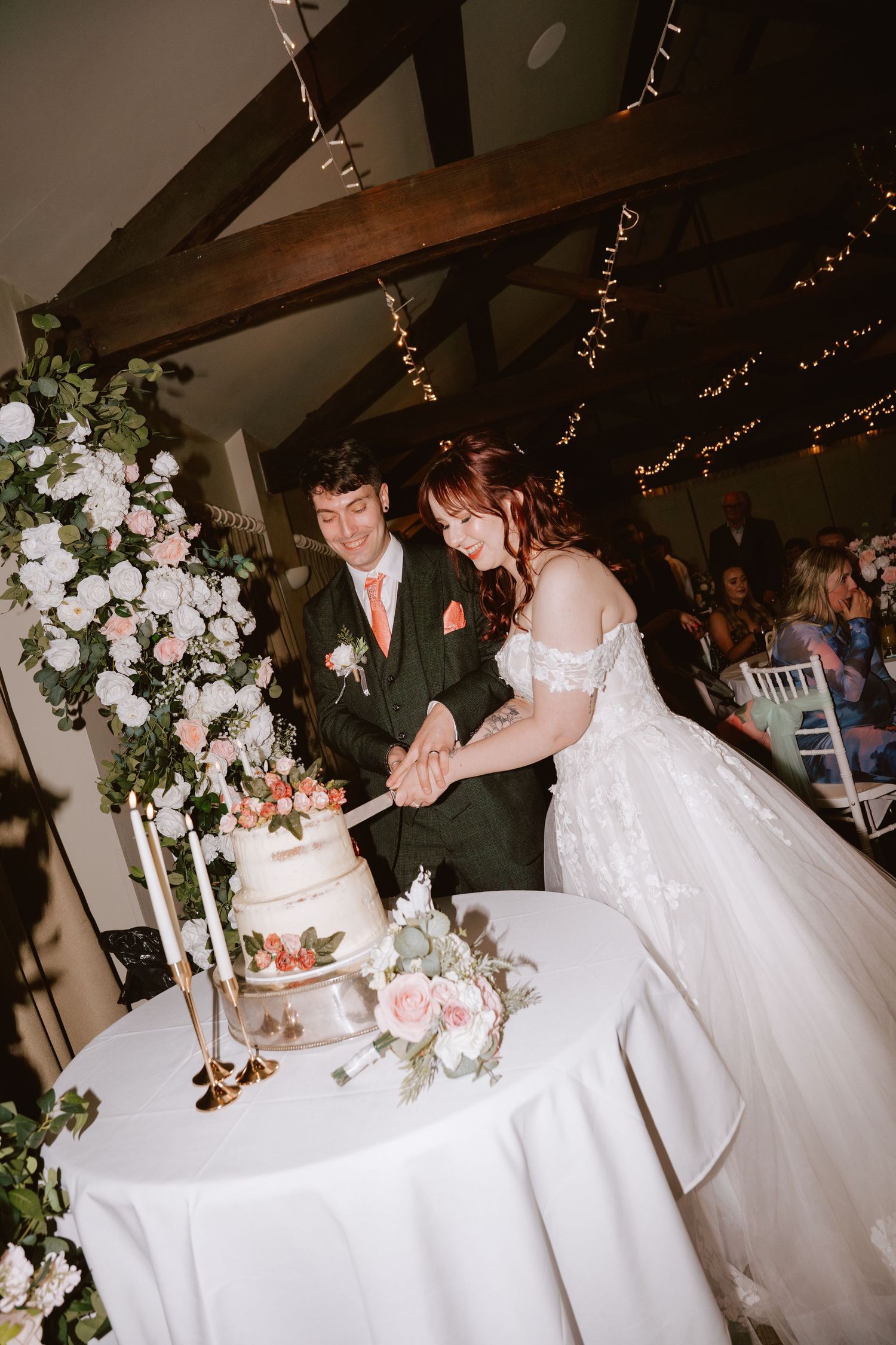 Bride and groom cutting their white tiered wedding cake at a rustic barn venue decorated with fairy lights and floral arrangements.