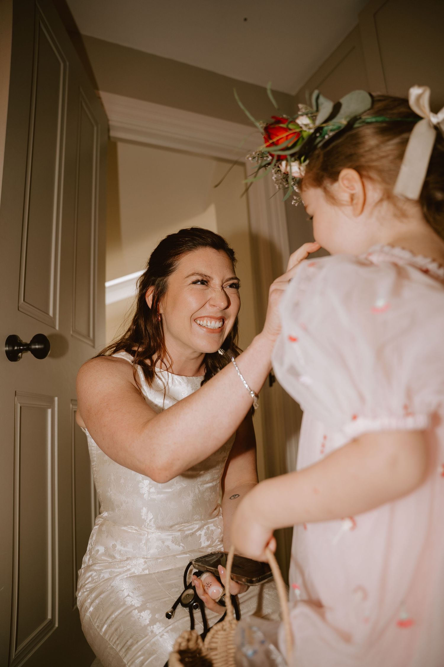 Bride in white gown smiling at flower girl wearing floral crown and pink dress during wedding preparations.