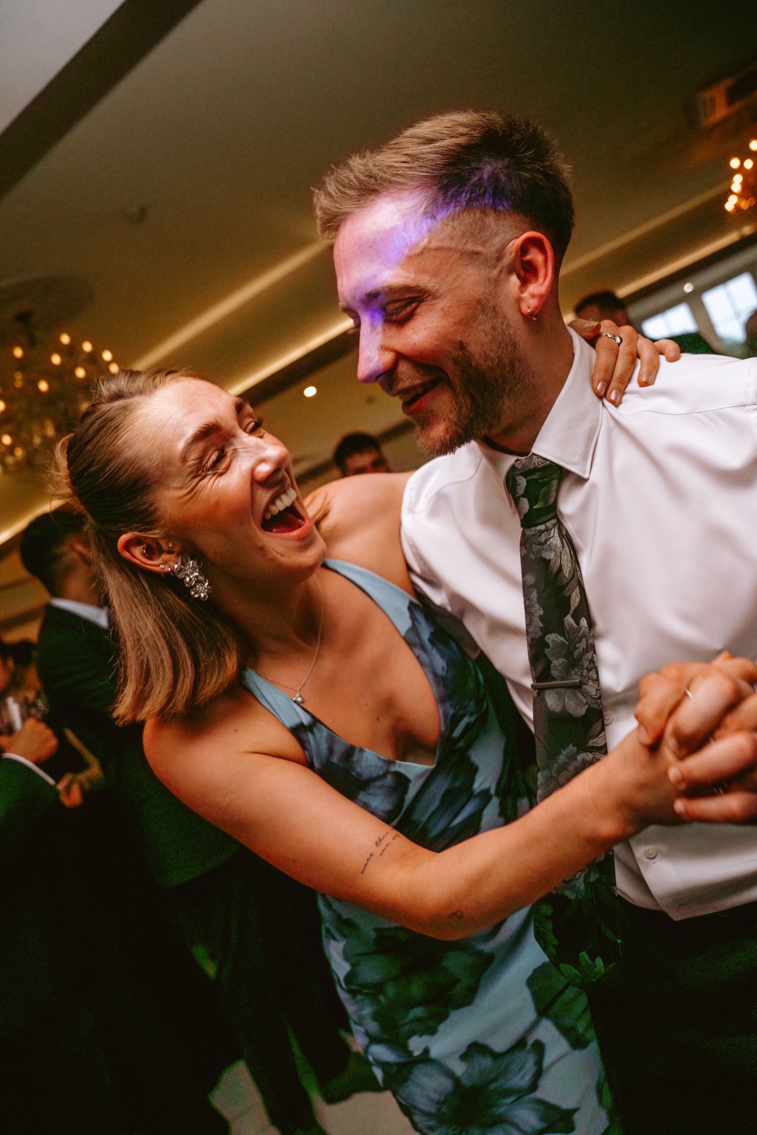 A joyful couple laughing and dancing together at a wedding reception in a warmly lit venue.