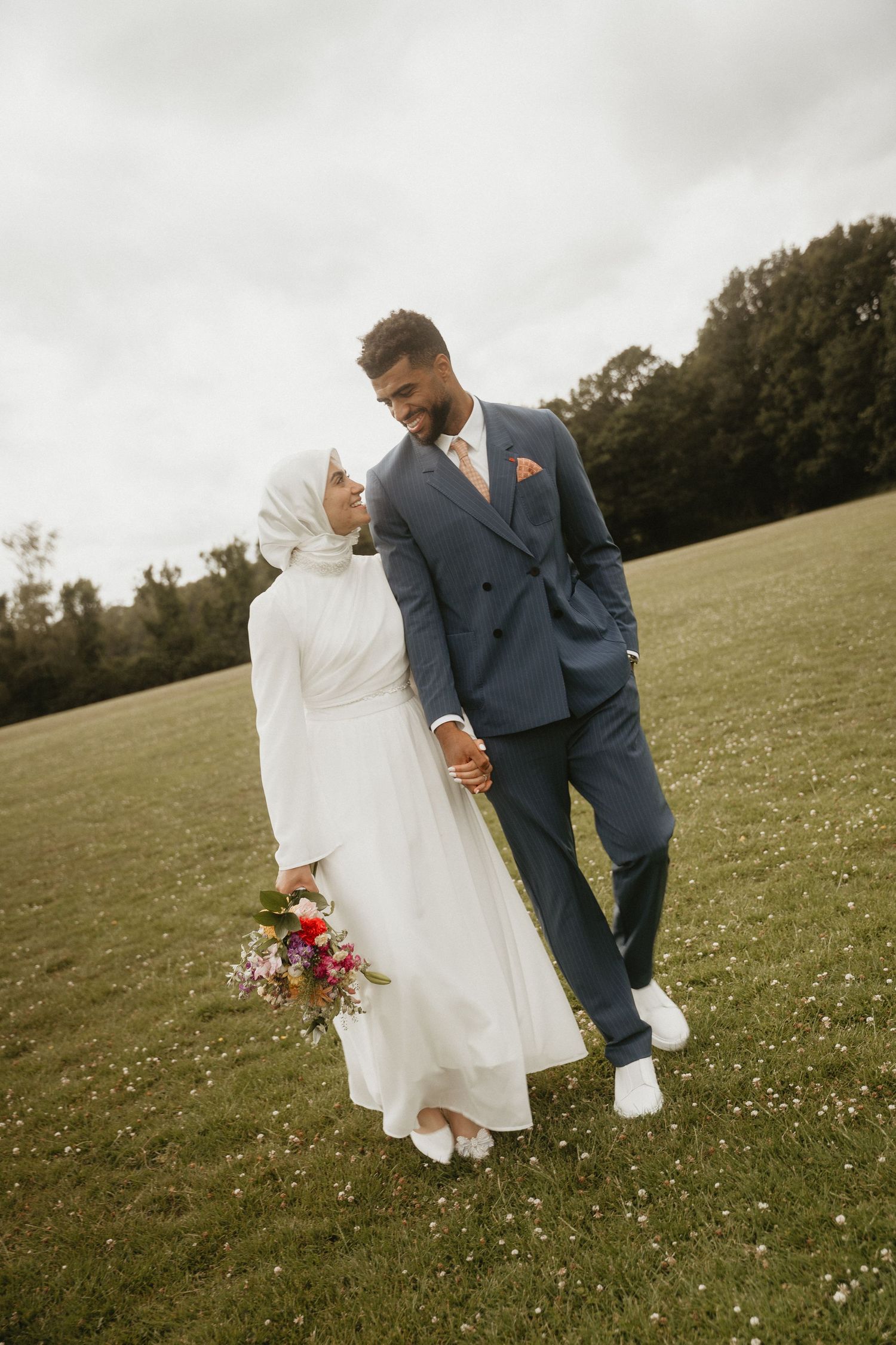 Artistic wedding photo of couple walking through meadow with vibrant bouquet.