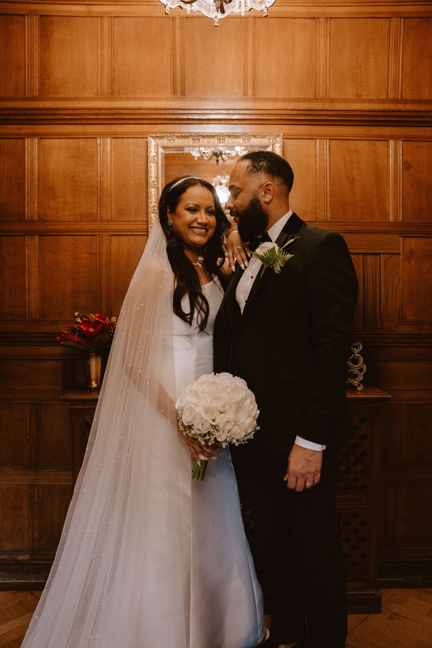 Bride in white gown holding bouquet smiles as groom in black tuxedo whispers to her in wood-paneled room.