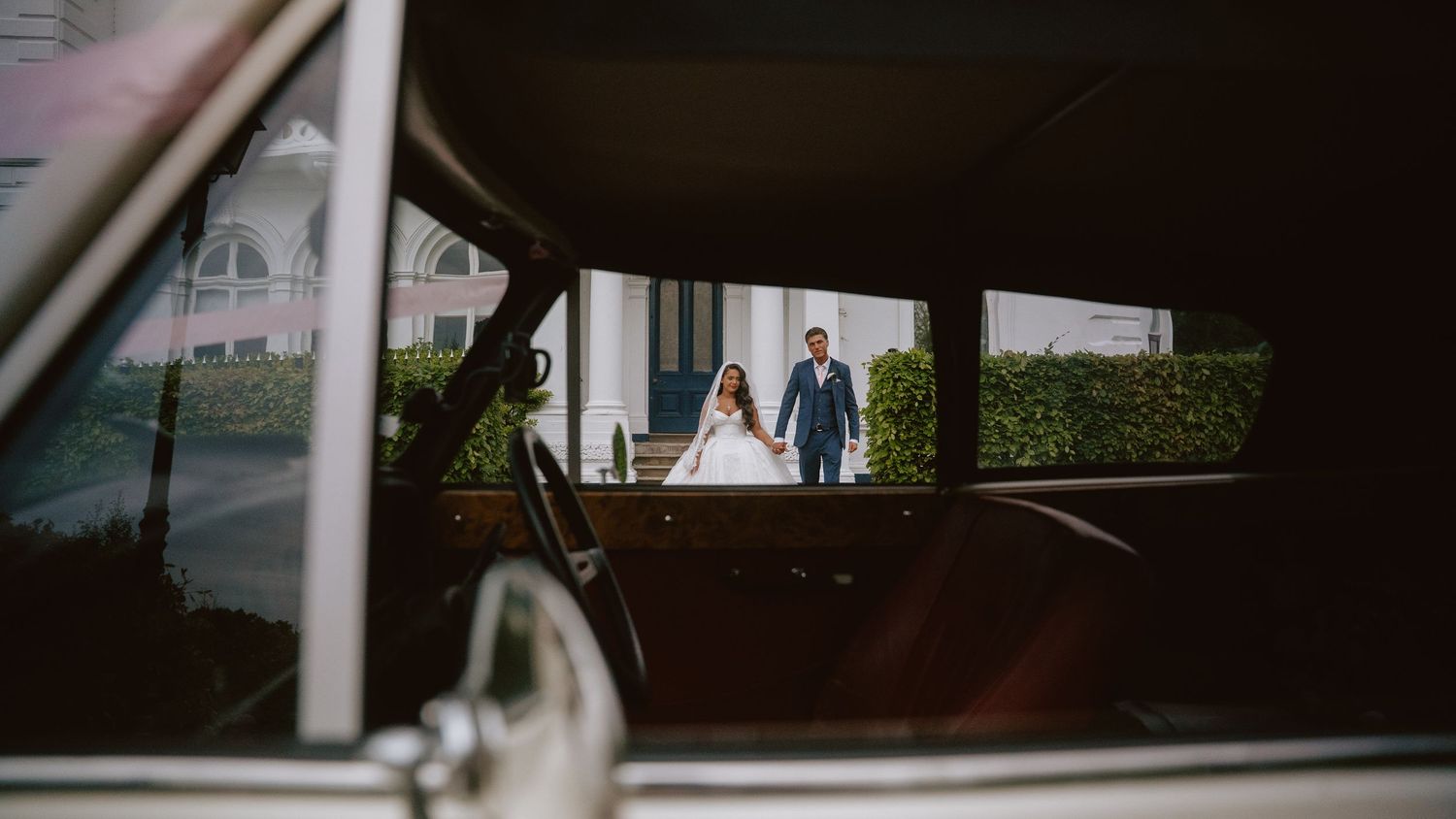 A bride and groom walk hand in hand, captured through the window of a vintage classic car at their wedding venue.