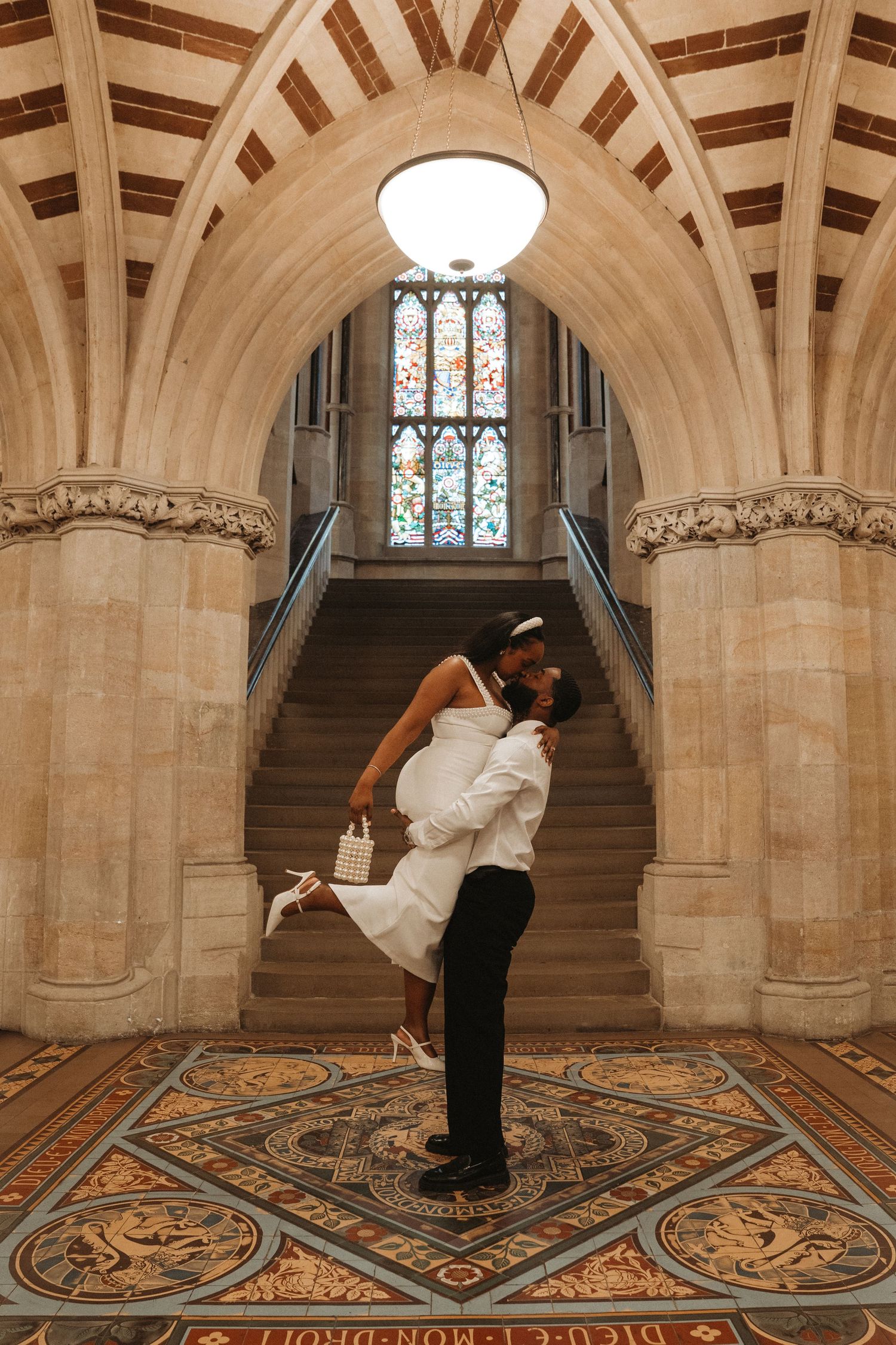A couple in wedding attire dips and kisses in a grand Gothic-style hall with ornate stone arches and patterned floor.