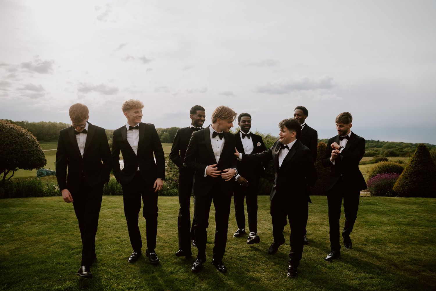 Seven groomsmen in black tuxedos with bow ties walking on a green lawn at an outdoor wedding celebration.