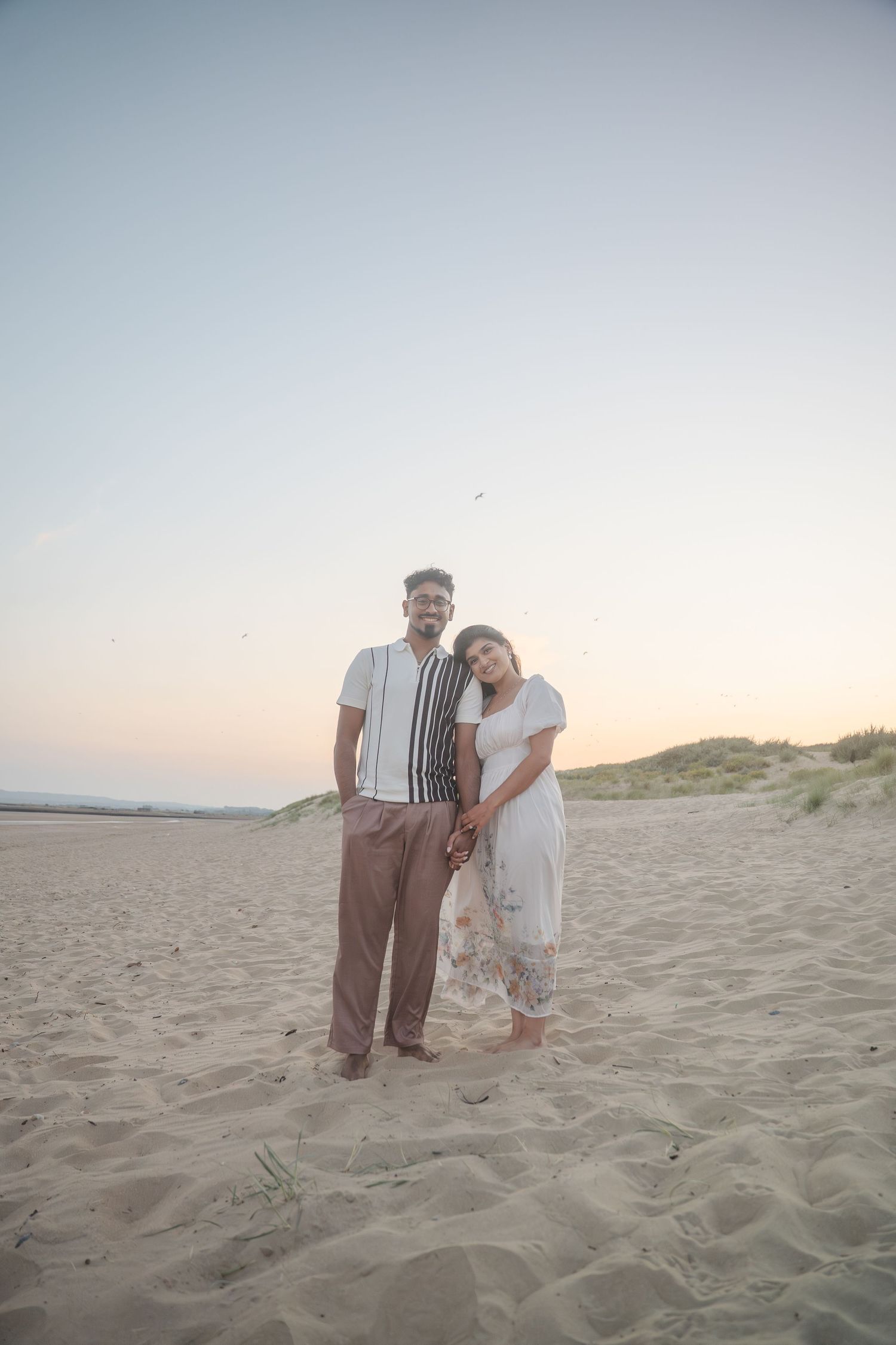 A couple poses together on a sandy beach at dusk wearing coordinated white and brown outfits.