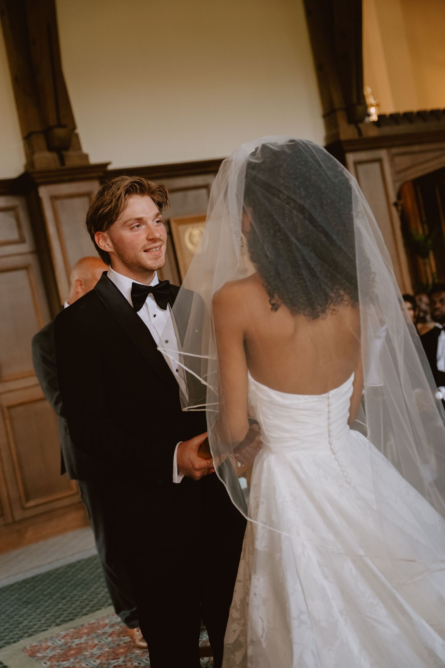 A groom in a black tuxedo smiles at his bride in a white strapless gown and veil during their wedding ceremony.