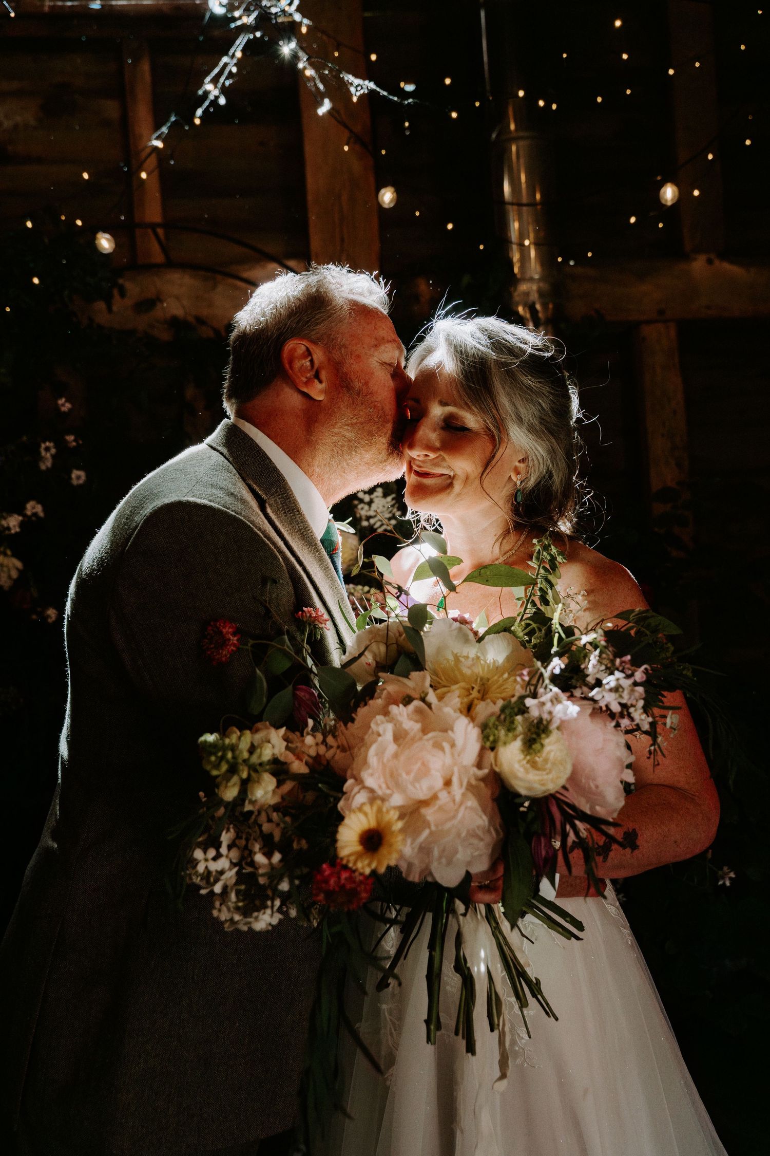 A bride and groom share a romantic kiss in a warmly lit barn wedding venue, bride holding a lush floral bouquet.