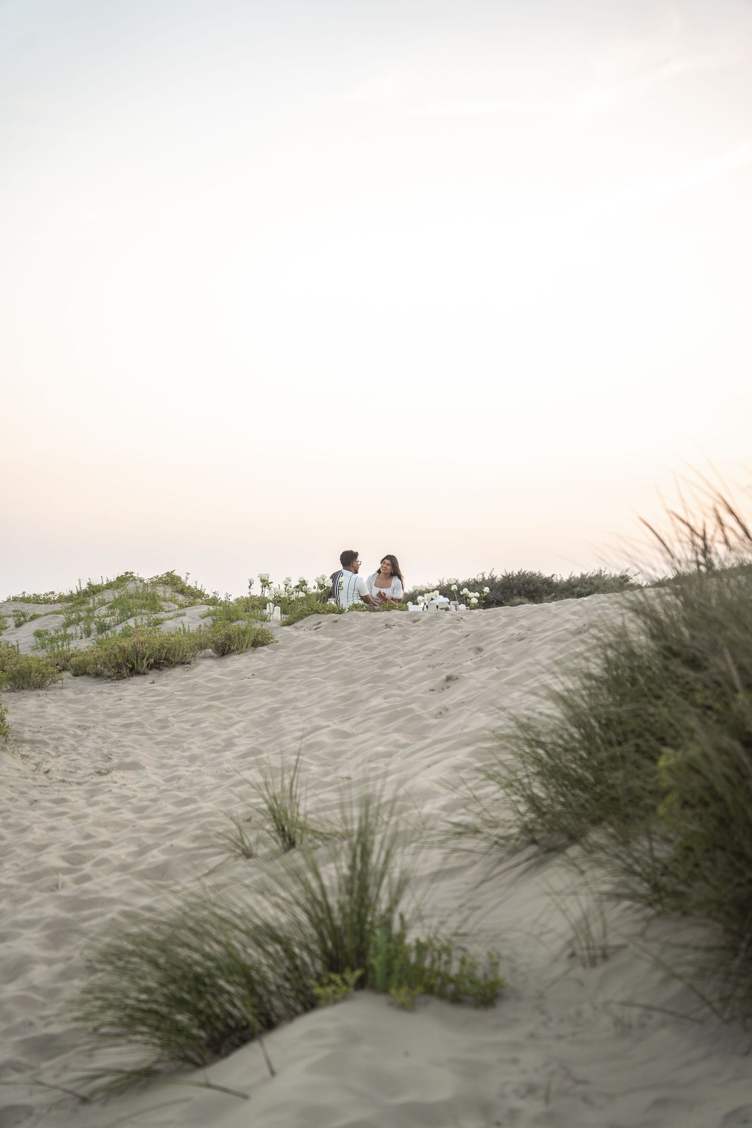 Couple sitting on sand dunes at sunset with beach grass in the foreground.