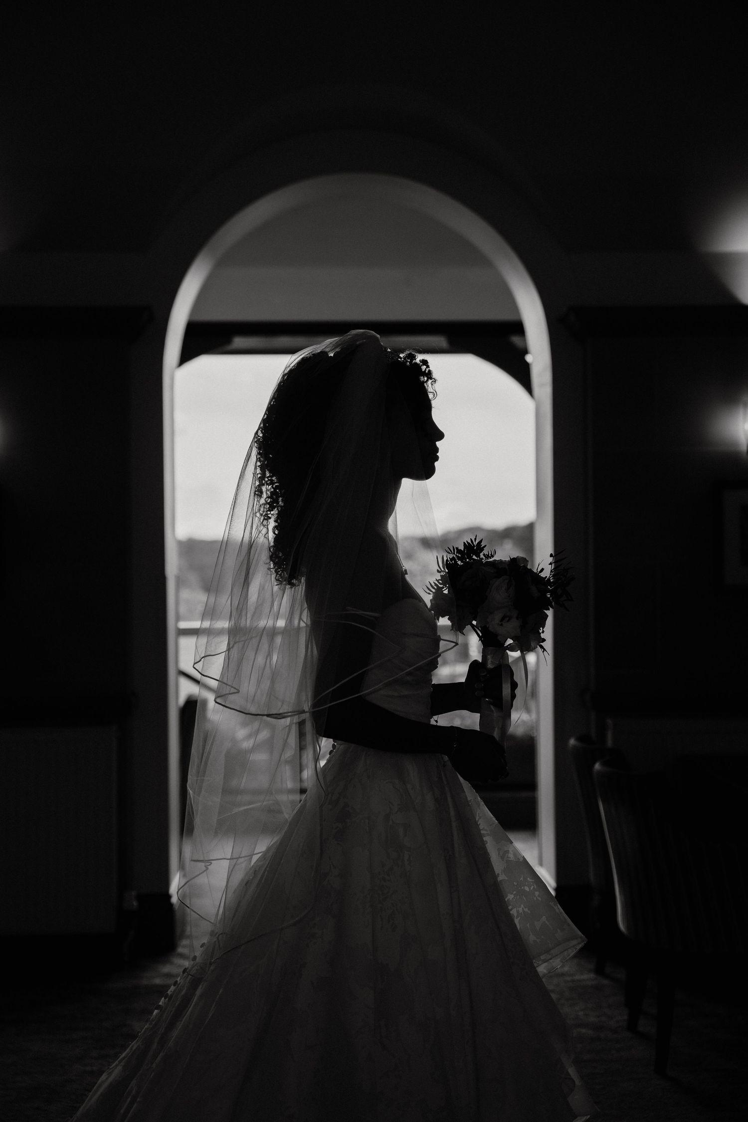 Black and white silhouette of a bride in wedding gown and veil holding bouquet, framed by an elegant arched doorway.