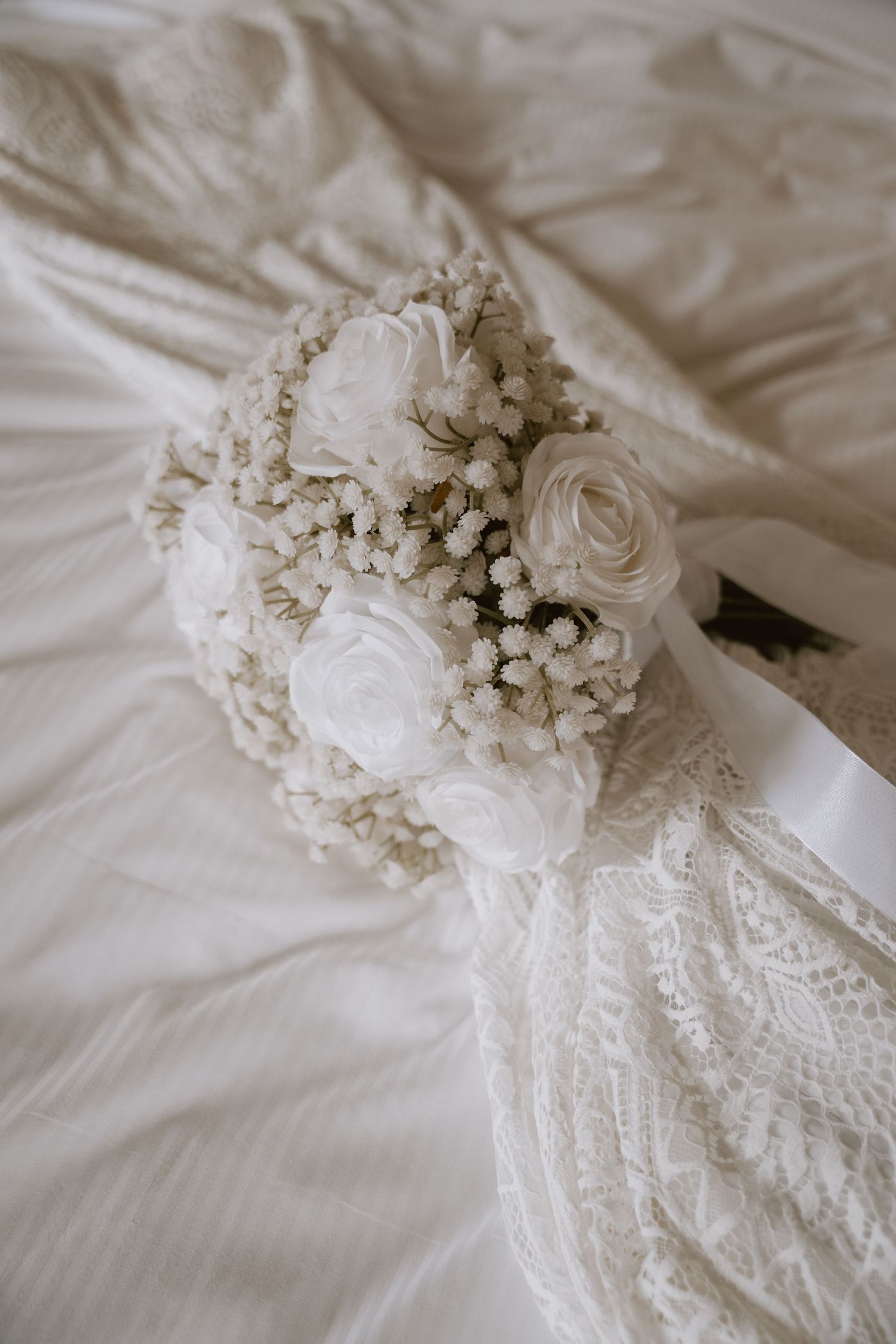 White roses and baby's breath bouquet rests on lace wedding dress fabric.