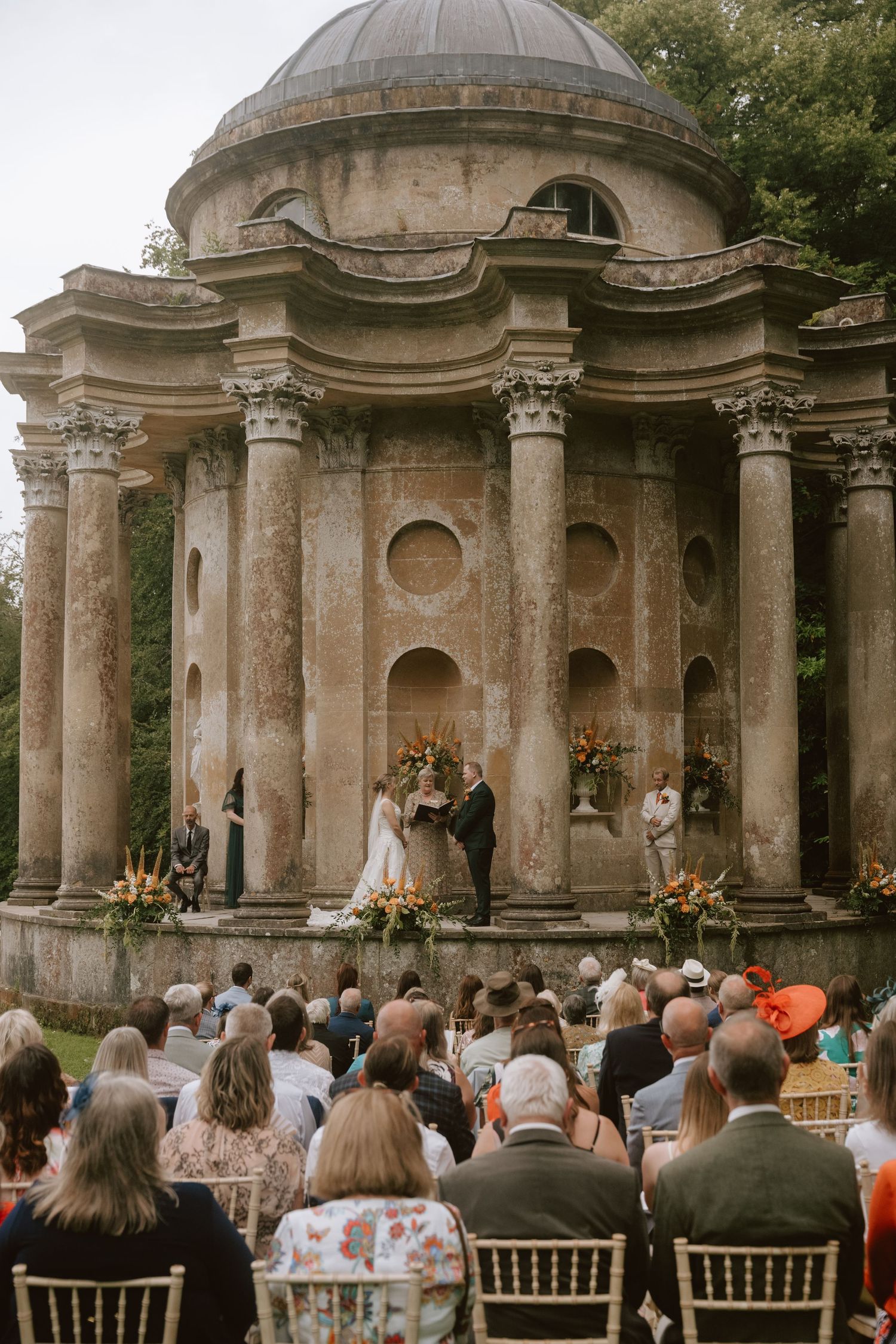 Wedding ceremony at a grand neoclassical temple with columns, guests seated outdoors, couple exchanging vows amid lush floral arrangements.
