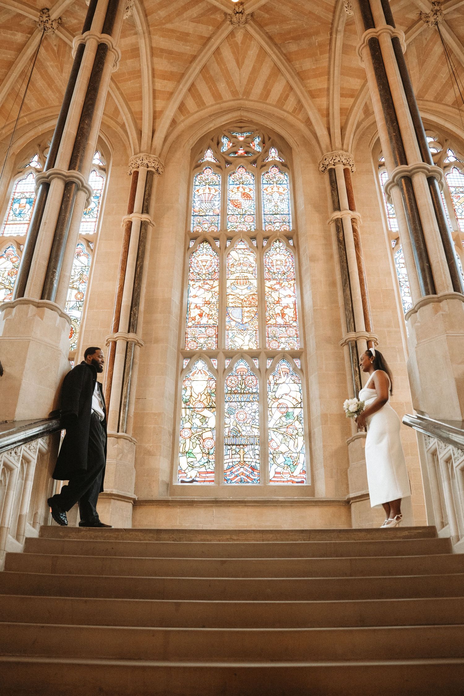 A couple walks up ornate stone stairs in a Gothic cathedral with stained glass windows and vaulted arches.
