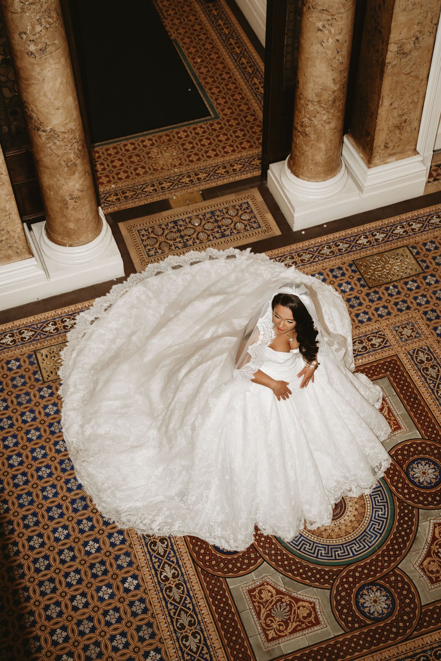 Aerial view of a bride in a voluminous white lace wedding gown seated on ornate tiled floor between grand columns.