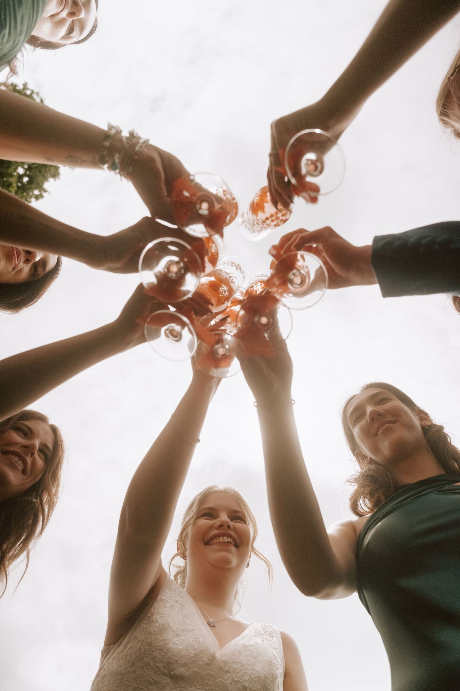 Bride and bridesmaids toasting with wine glasses viewed from below at a wedding celebration outdoors.