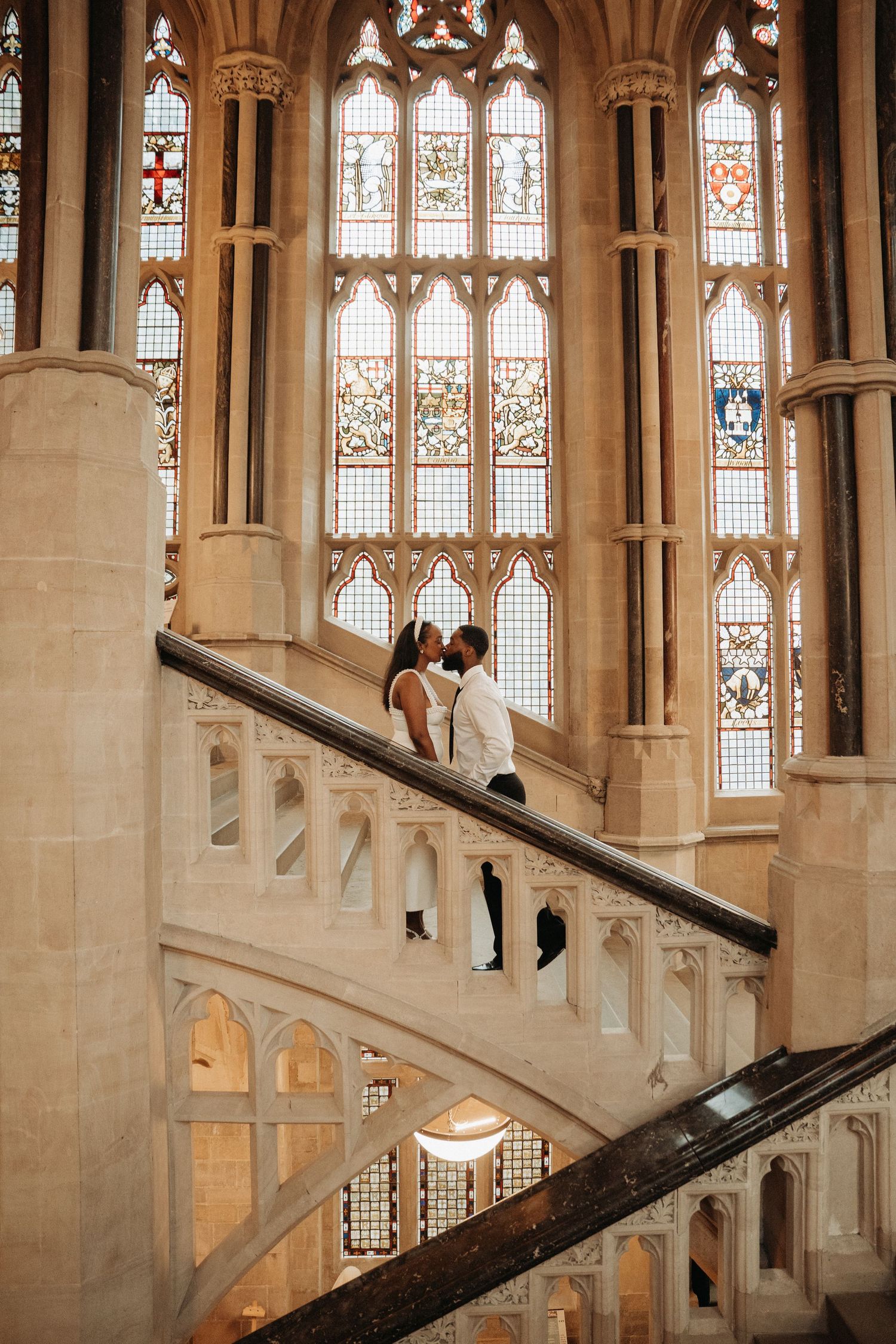 Couple sharing a romantic moment on an ornate spiral staircase inside a grand Gothic cathedral with stained glass windows.