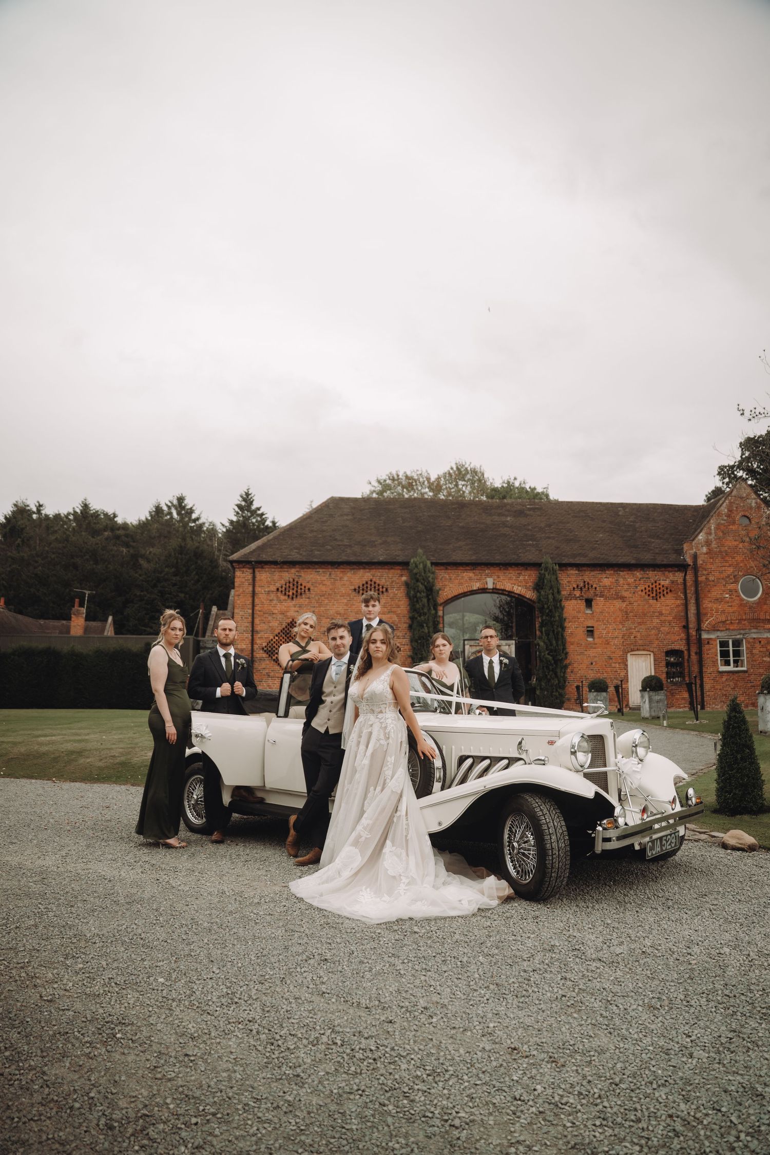 Wedding party posing with vintage white car outside brick venue on overcast day.