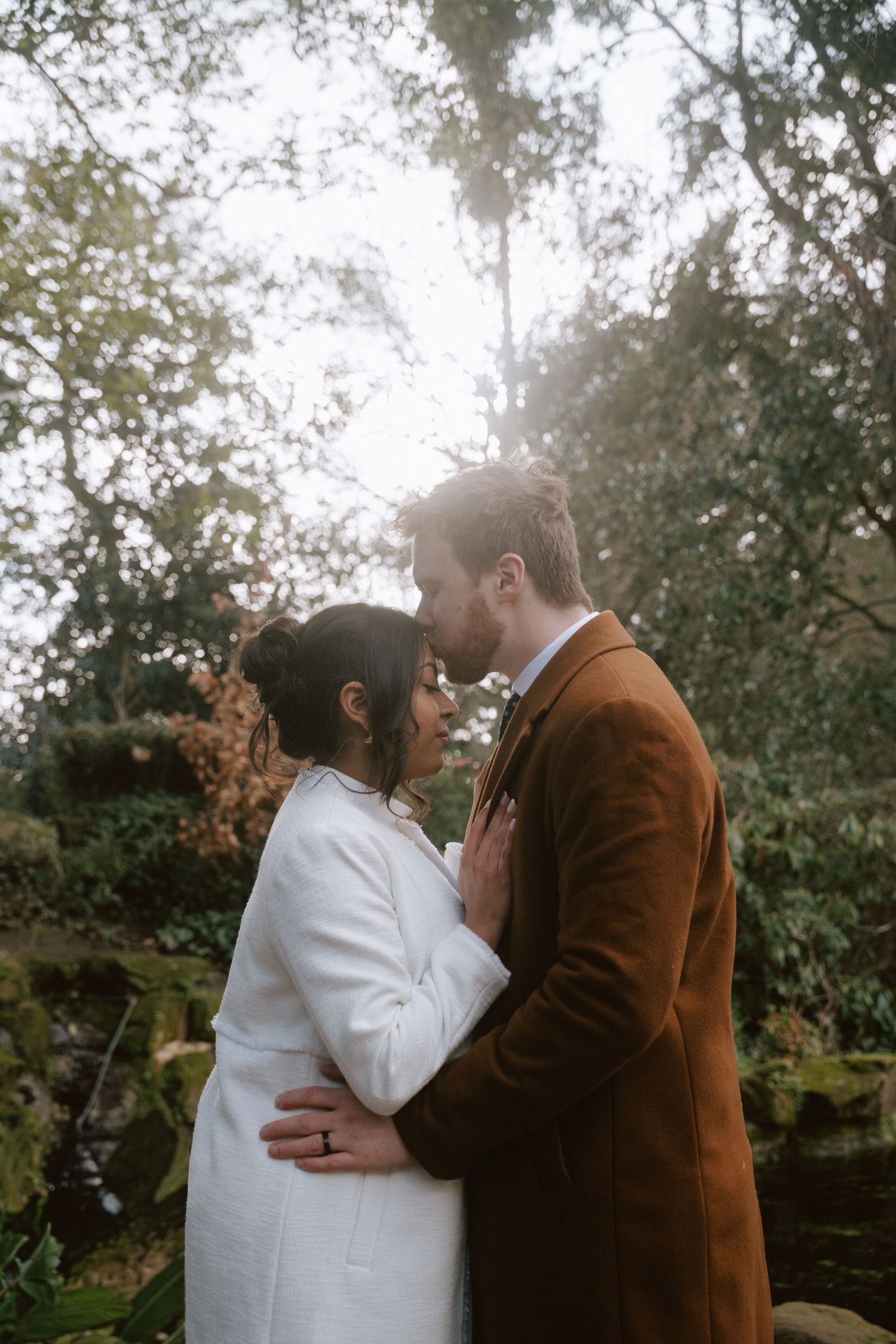A couple shares an intimate moment in a garden setting wearing contrasting brown and white outfits.