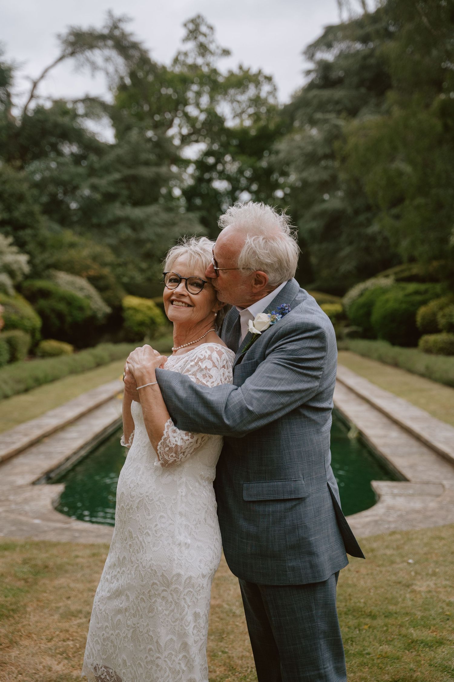 Elderly couple in wedding attire embracing by a garden pool, groom kissing bride's cheek as she smiles.