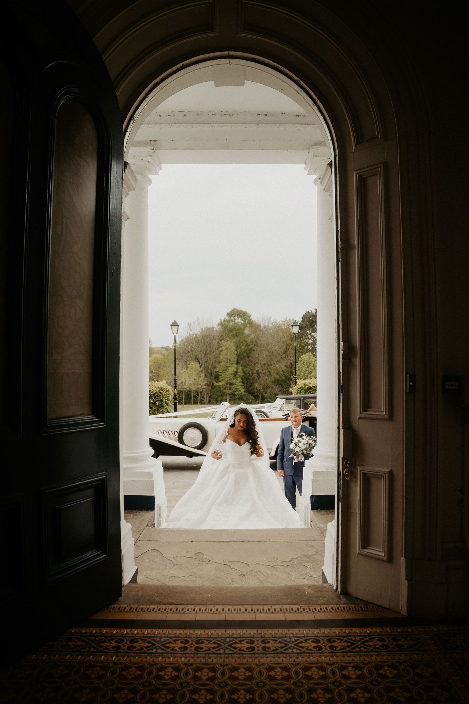 Bride in a white ball gown entering elegant venue doors with escort, lush greenery visible outside.