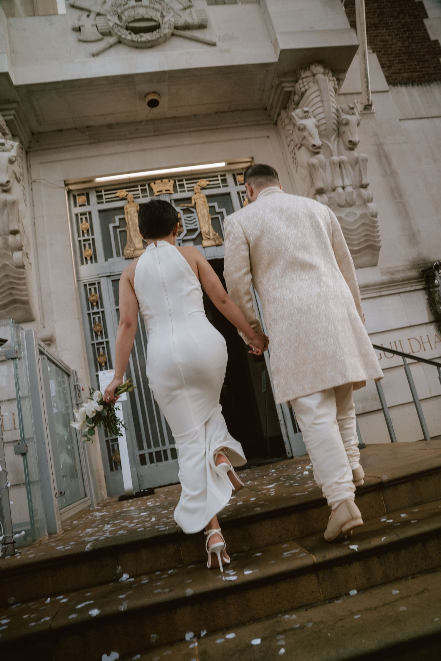 Two people in white formal attire walk together along an ornate building exterior.