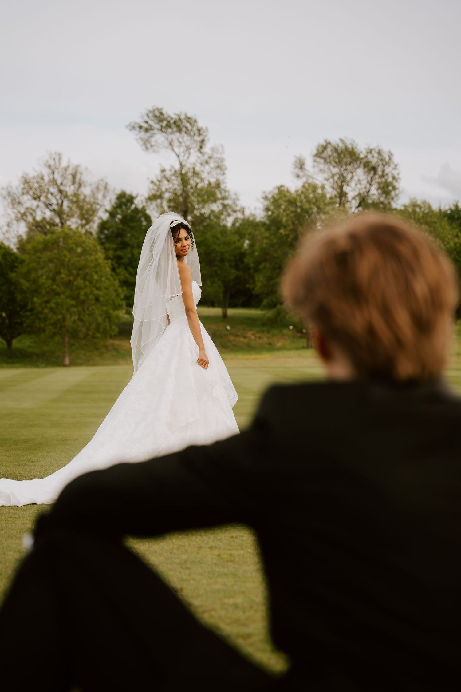 Bride in white gown and veil stands on green lawn as groom watches from foreground at outdoor wedding.