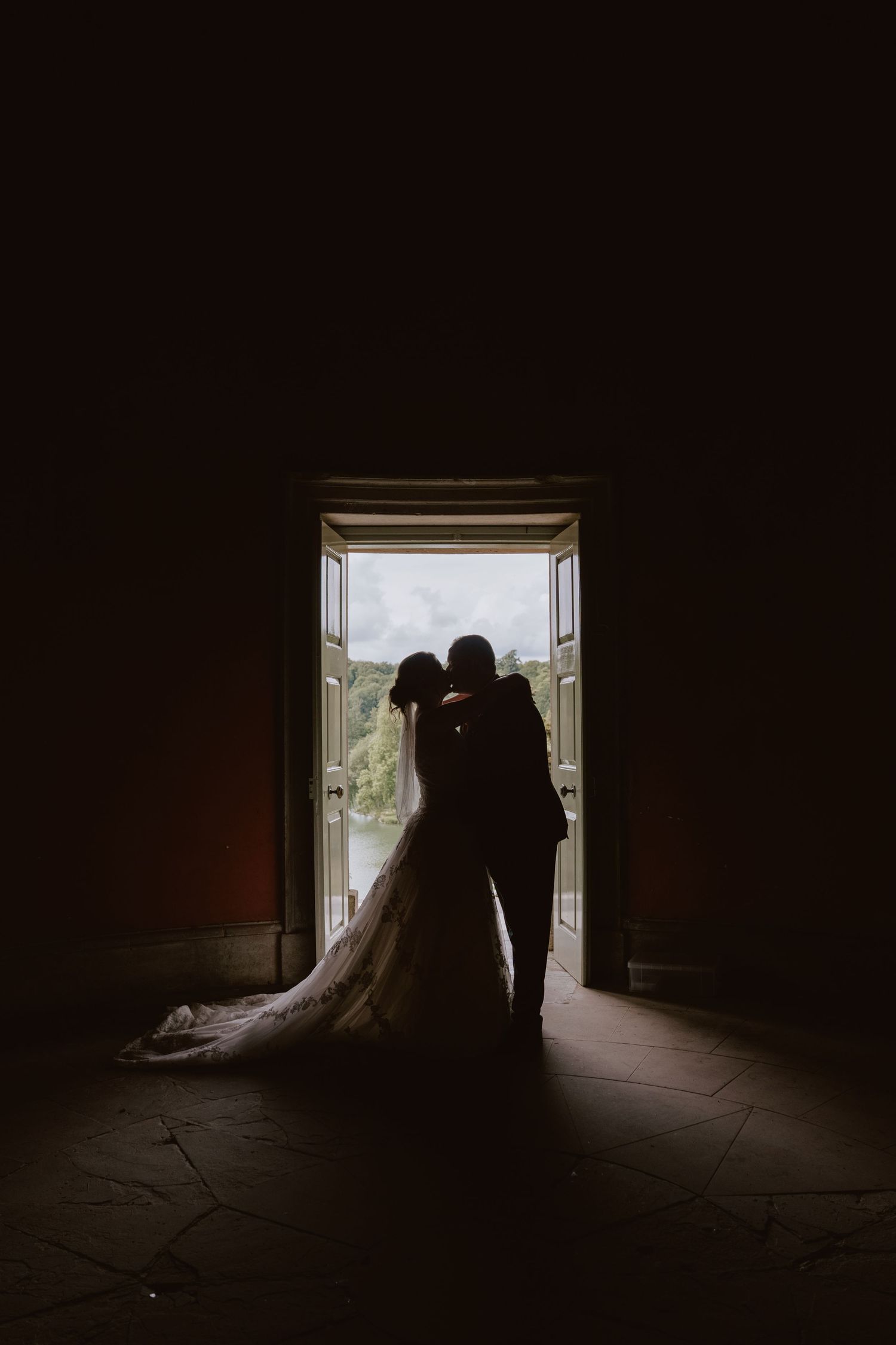 Silhouette of bride and groom kissing in doorway with flowing wedding dress train, backlit by natural light.