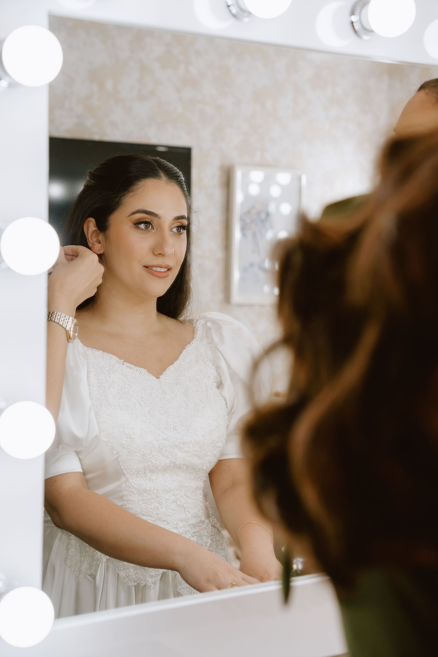 Bride in white lace dress adjusting earring before Hollywood vanity mirror with bulb lights.