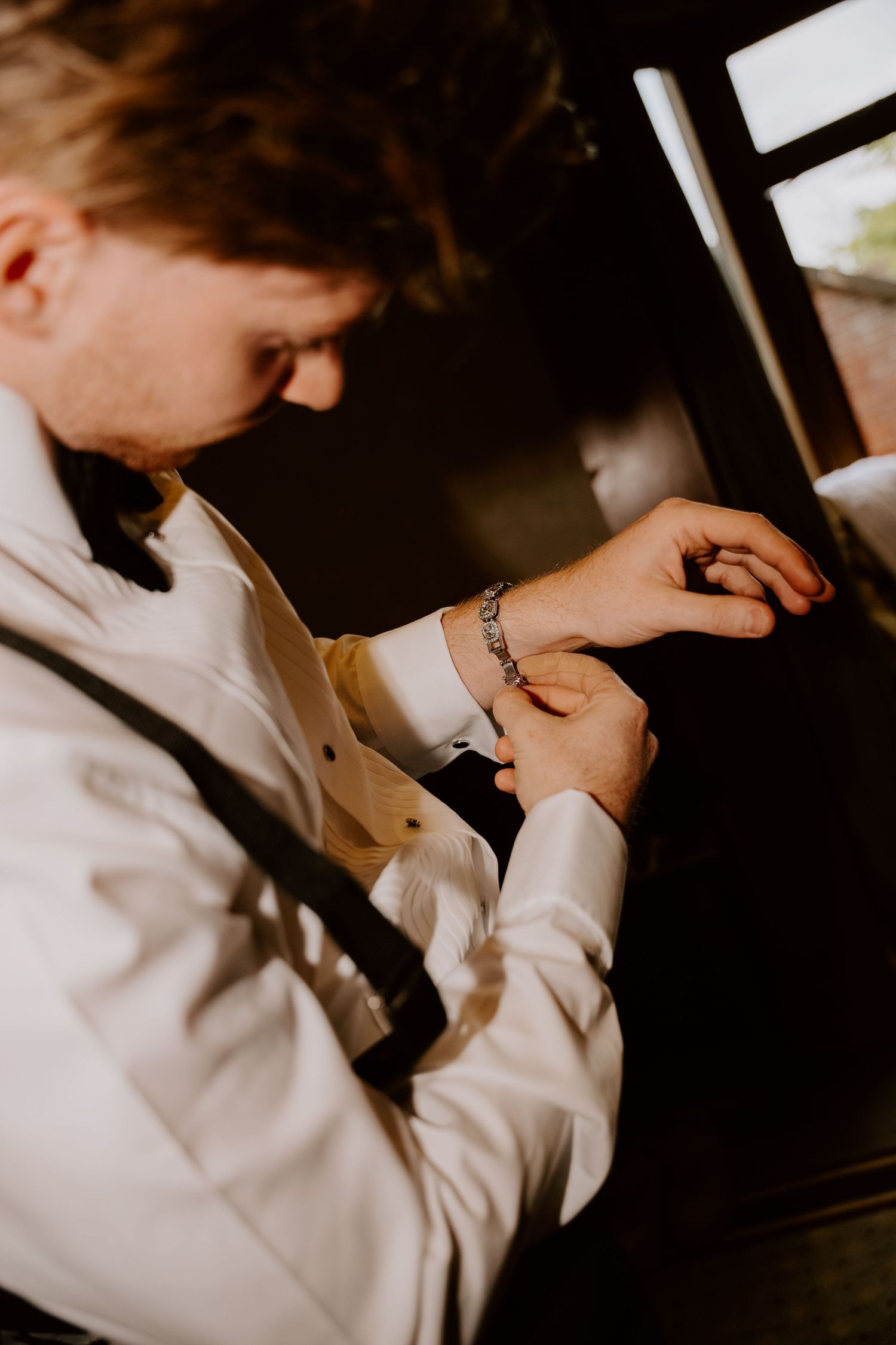 Groom in white dress shirt and suspenders fastening his cufflinks while preparing for his wedding day.