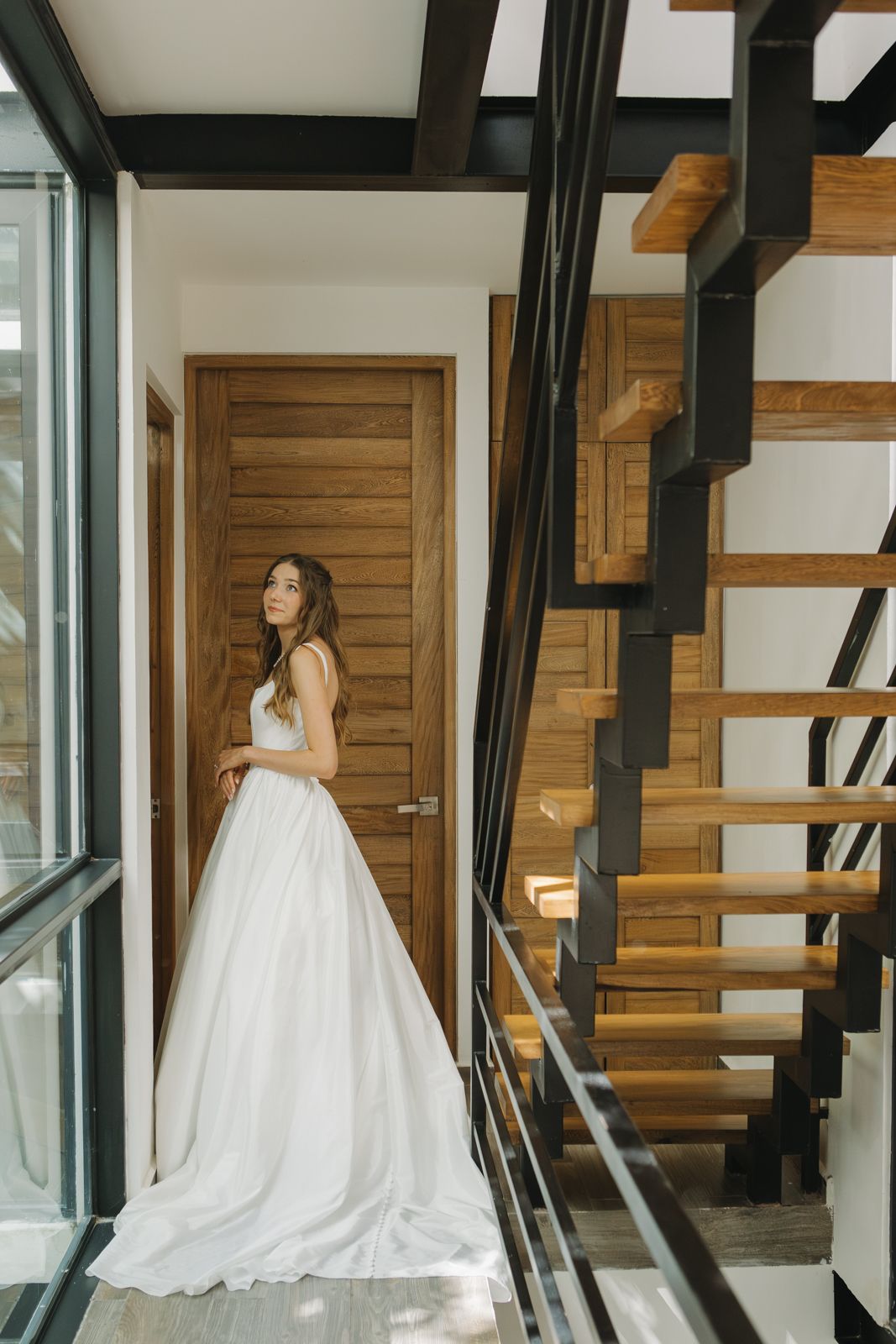 A bride standing in a hallway next to a window on her wedding morning.