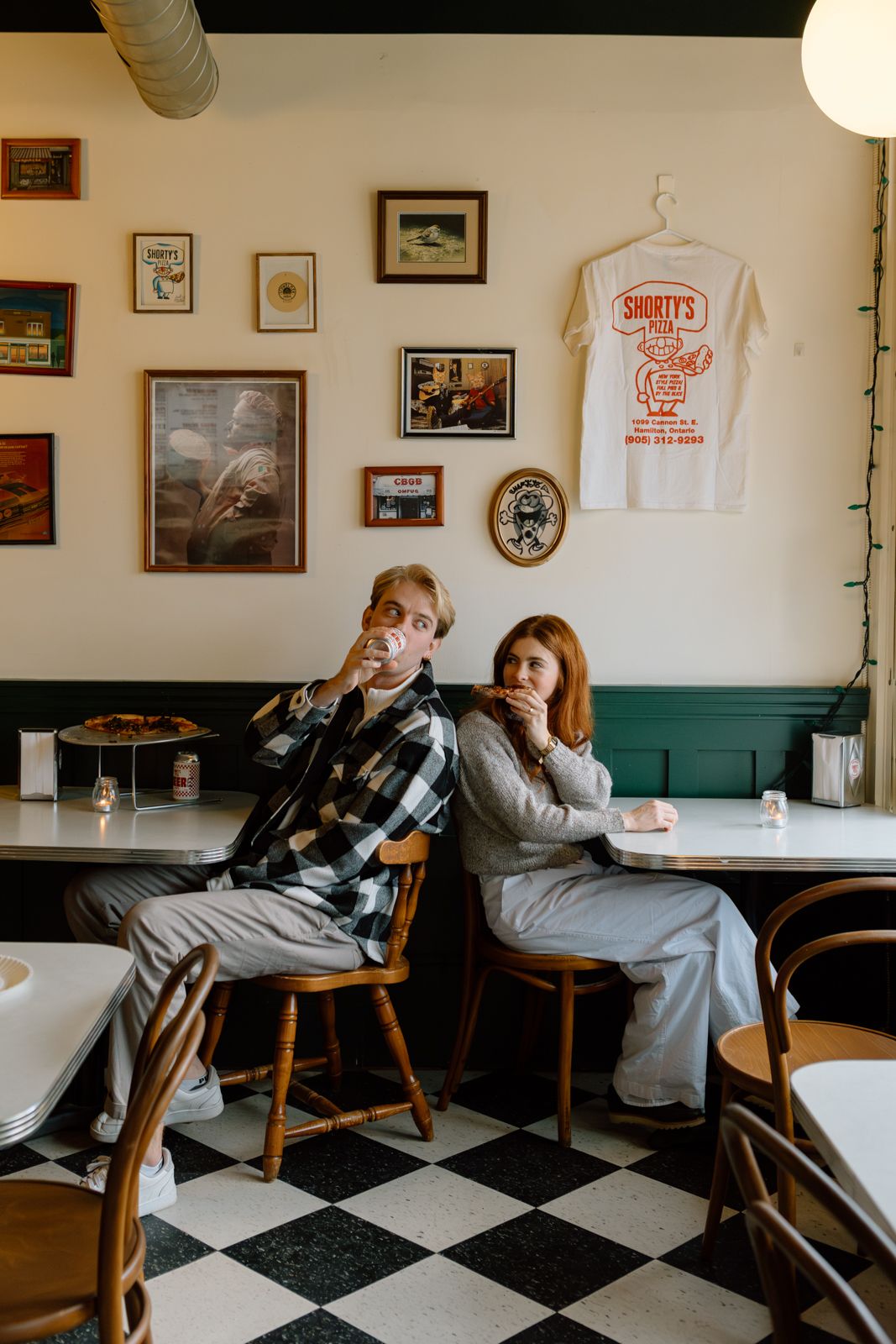 An engaged couple sitting back-to-back in chairs at a pizza parlour next to a gallery wall.